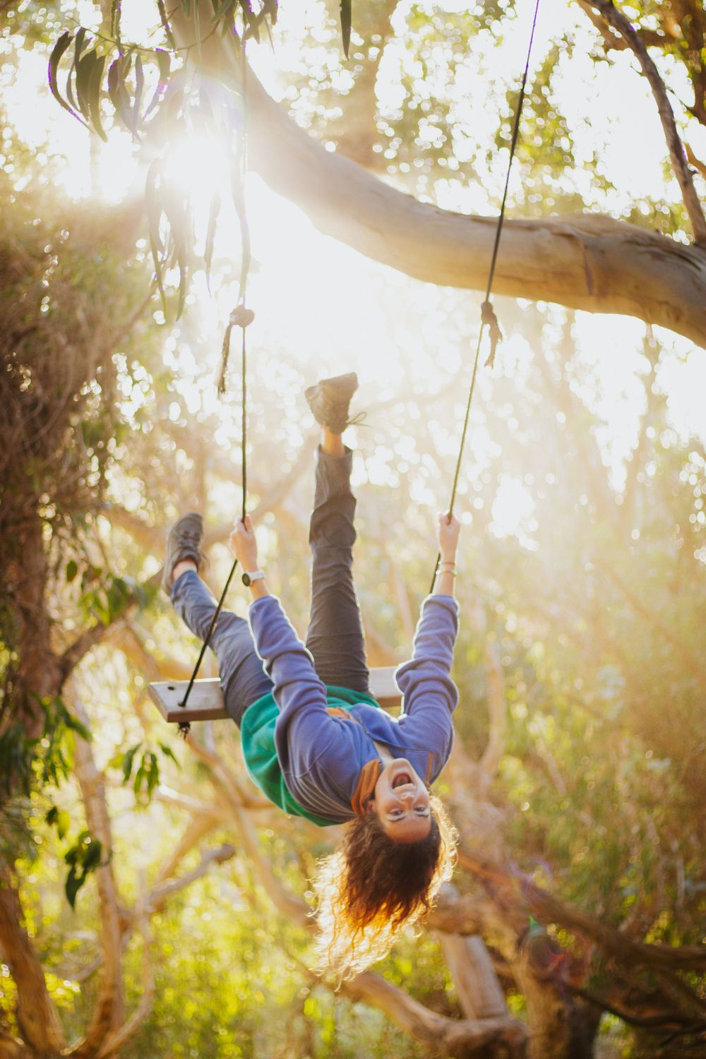 Person playfully hanging upside down from tree swing in sunlit forest setting.