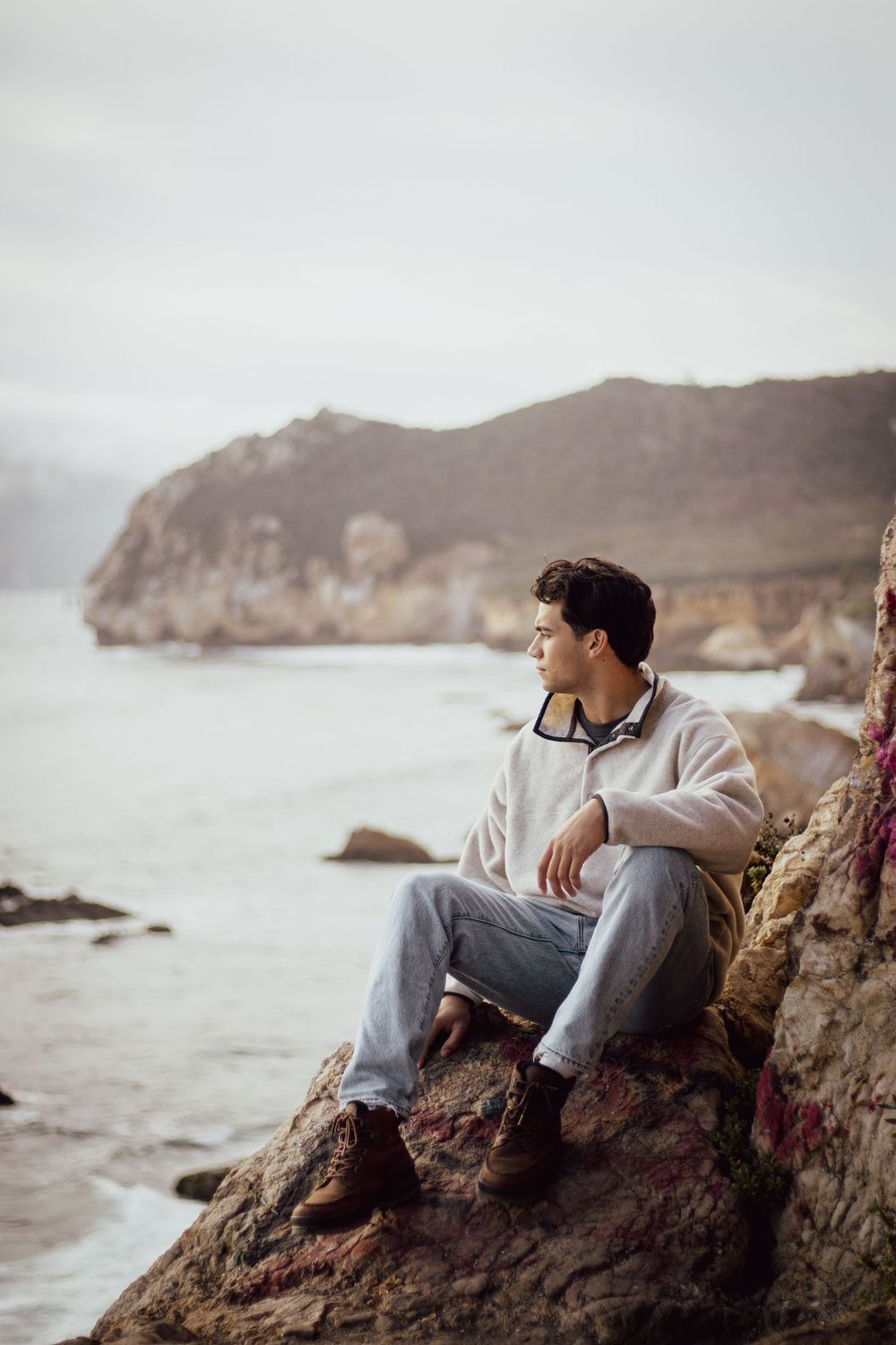 Figure sits on coastal rocks contemplating scenic ocean vista on foggy day.