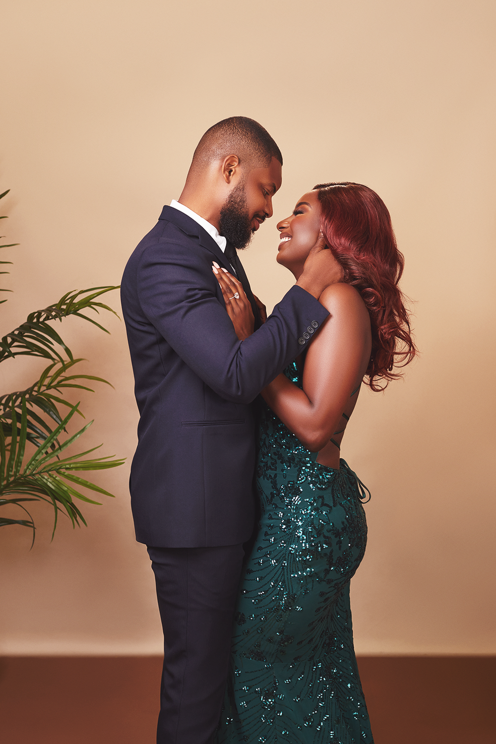A romantic couple embraces while dressed formally in a navy suit and sparkling emerald gown against a neutral backdrop.