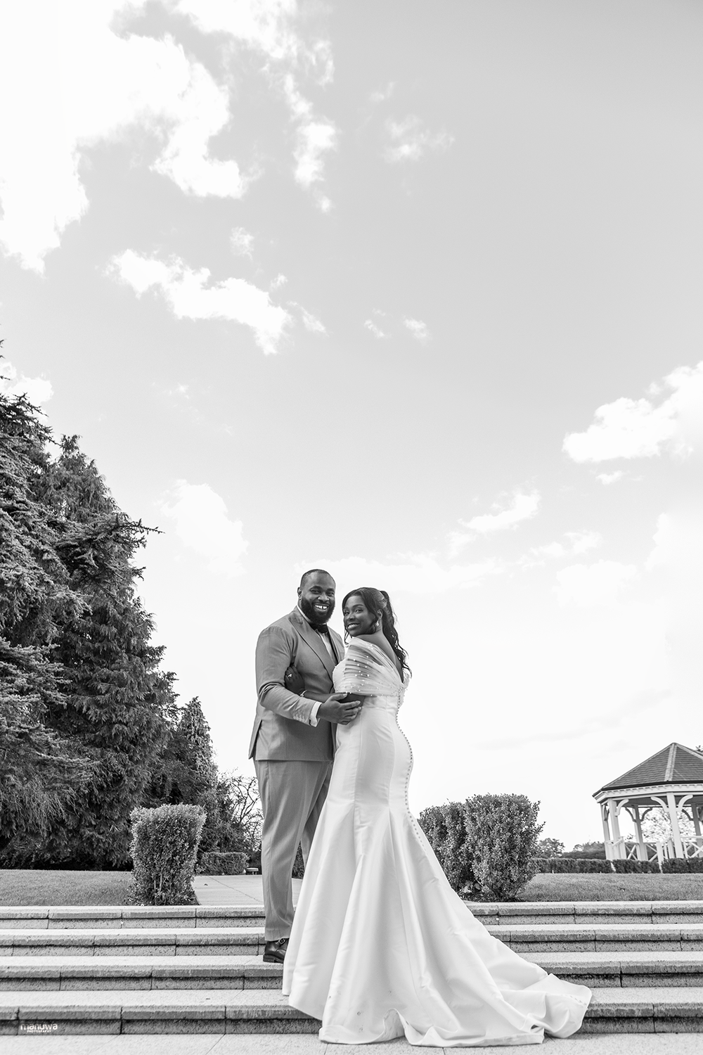 A black and white portrait of newlyweds standing on stone steps with a gazebo in the background.