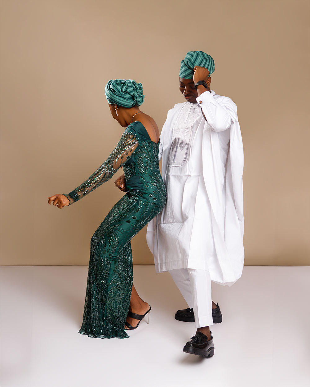 Two people in traditional Nigerian attire, one wearing a sparkly green dress and headwrap, the other in white agbada dancing.