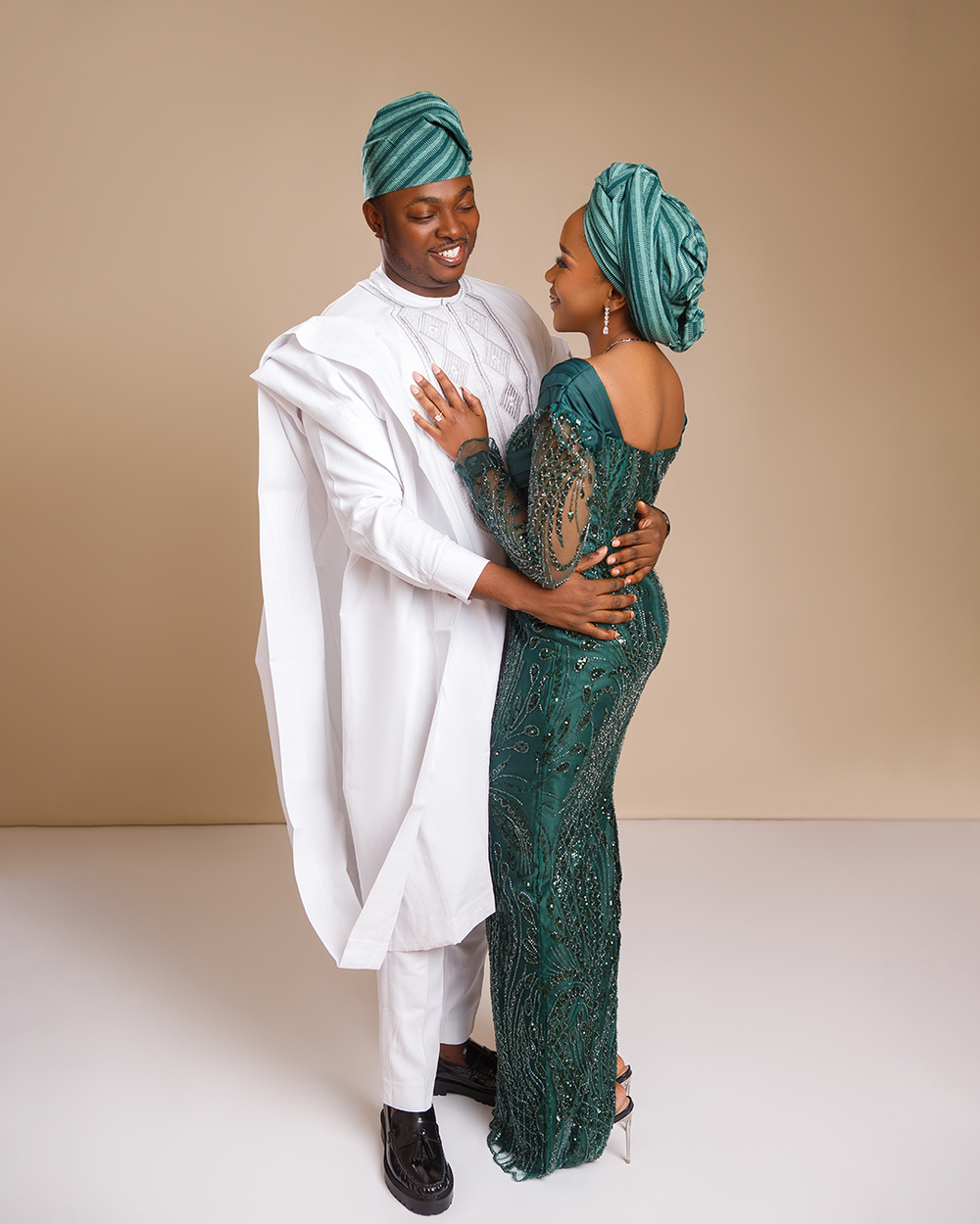 Couple in coordinated traditional African attire with teal headwraps and white agbada poses elegantly against beige backdrop.