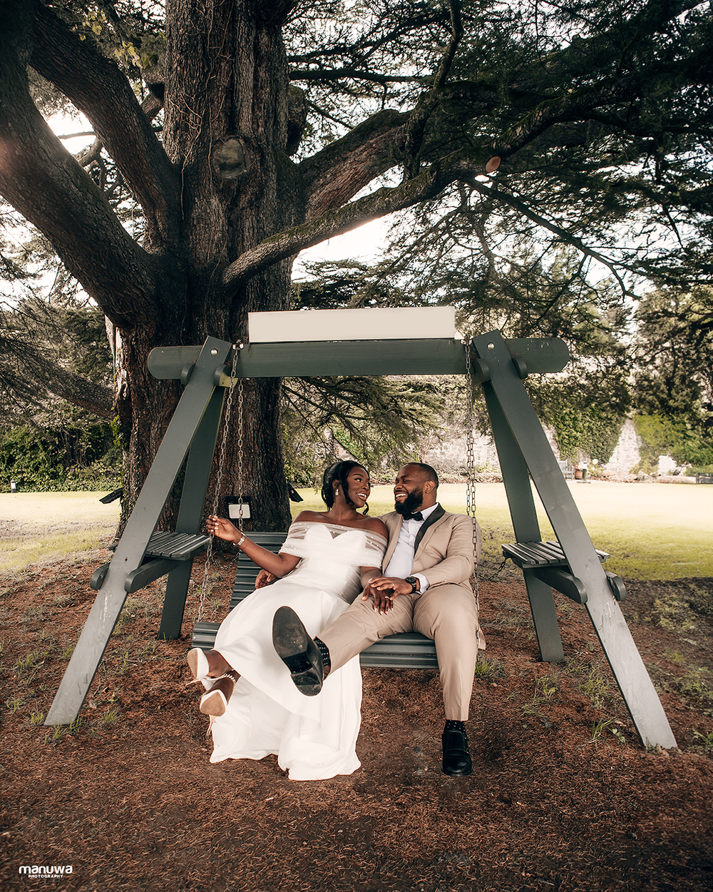 A couple shares a romantic moment on a white swing beneath a large tree in elegant cream-colored attire.