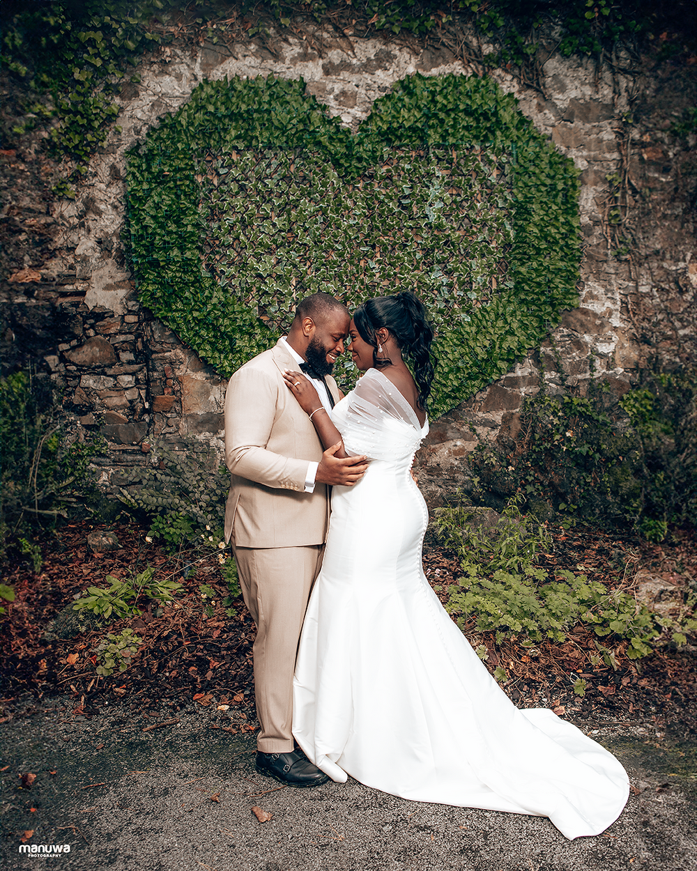 A couple embraces in front of a heart-shaped ivy wall, with the bride in white and groom in beige.