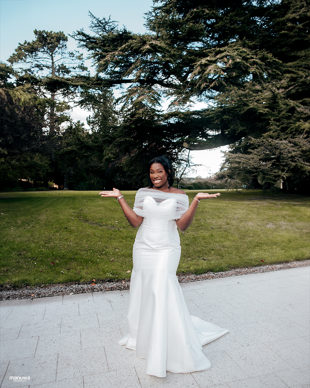A bride poses joyfully in a fitted white wedding gown against a backdrop of evergreen trees.
