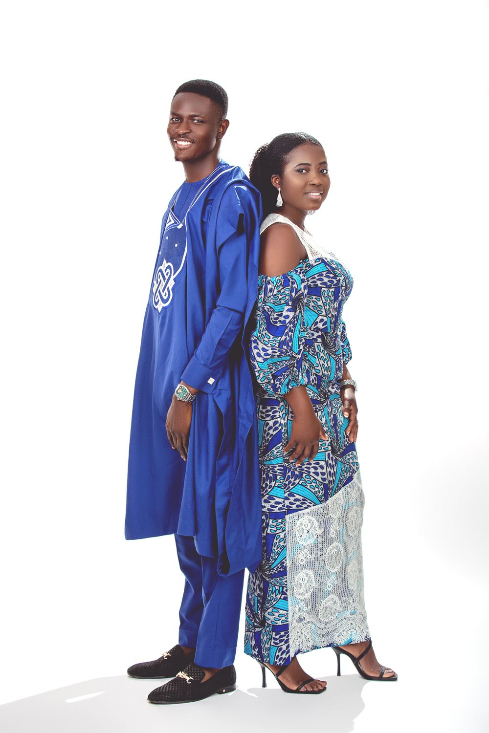 Two people pose back-to-back in traditional African attire, wearing blue and patterned garments against a white background.