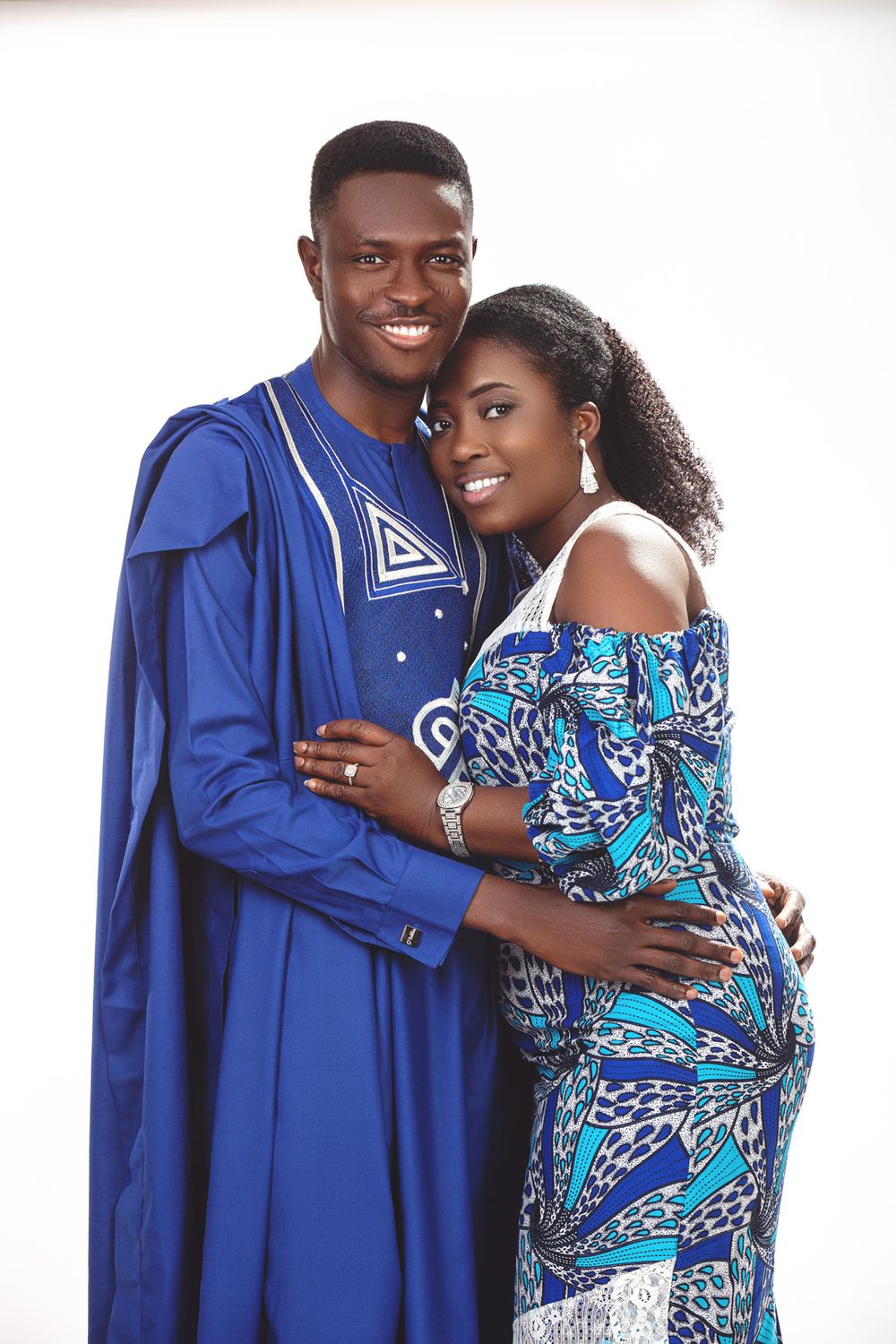 A couple in traditional African attire embrace and smile, wearing blue agbada and printed ankara fabric dress.