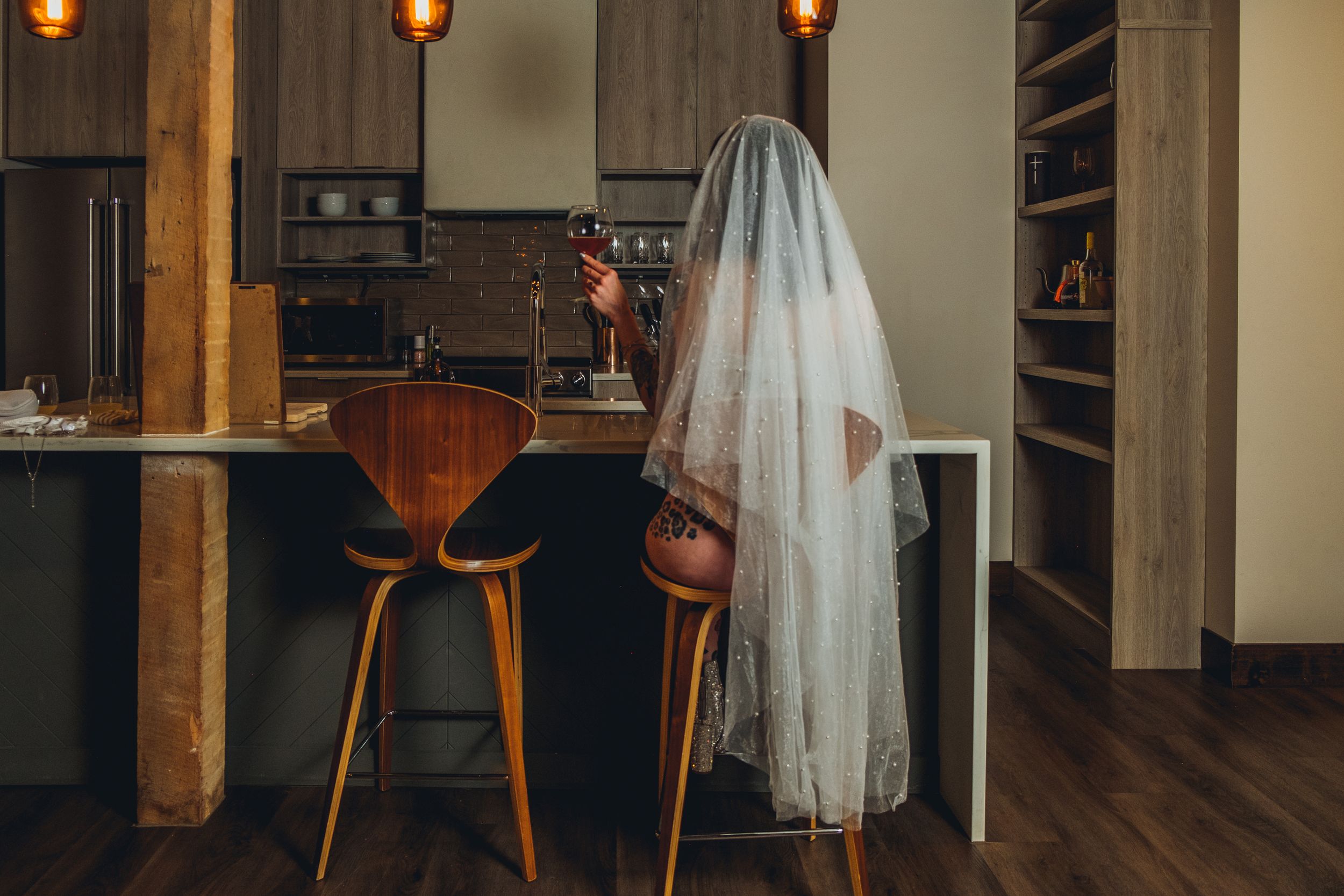 A veil-draped figure stands near wooden bar stools in a modern kitchen with warm lighting and hardwood floors. bridal boudoir
