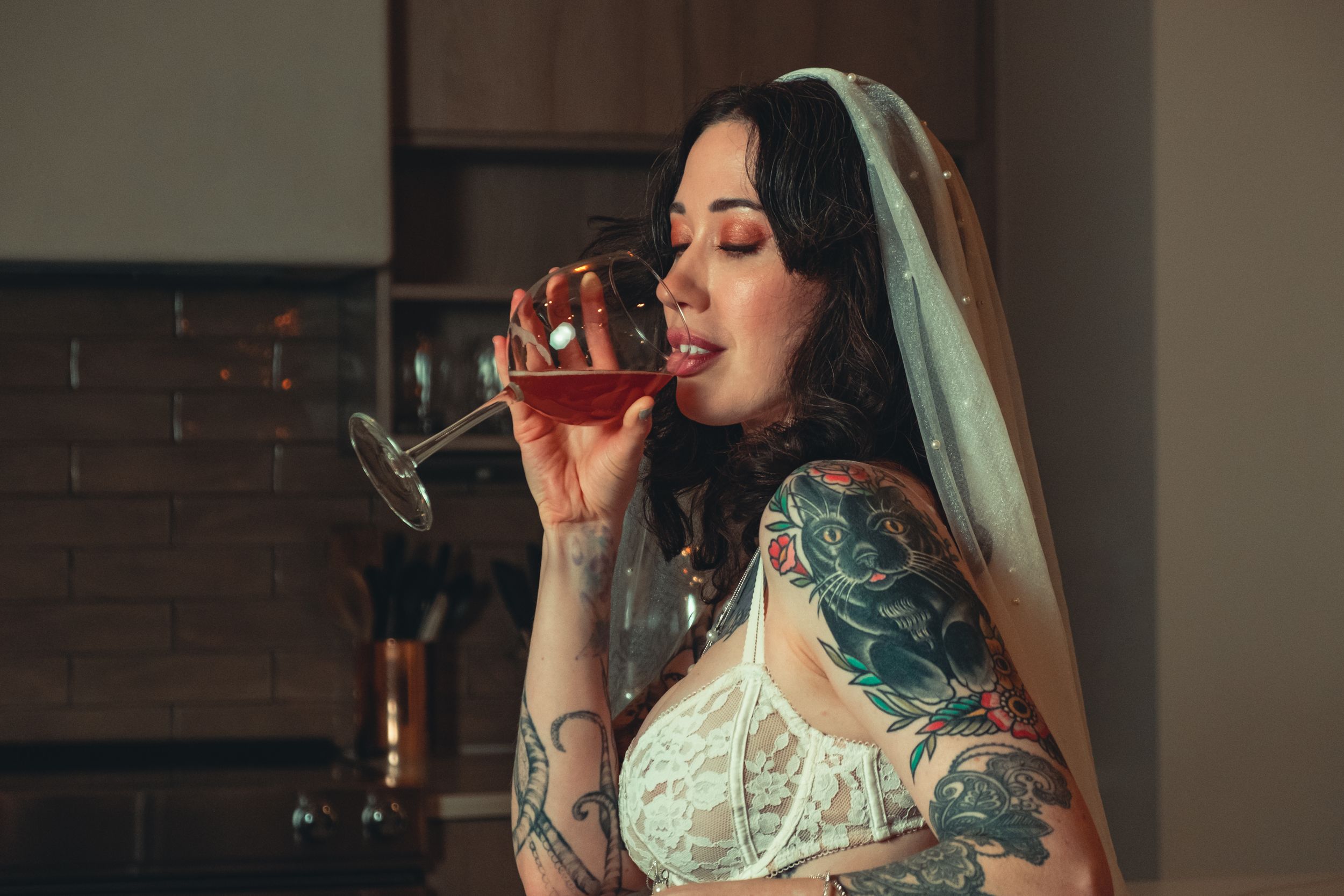 Artistic shot of someone in white lace sipping wine in a dimly lit kitchen setting. bridal boudoir