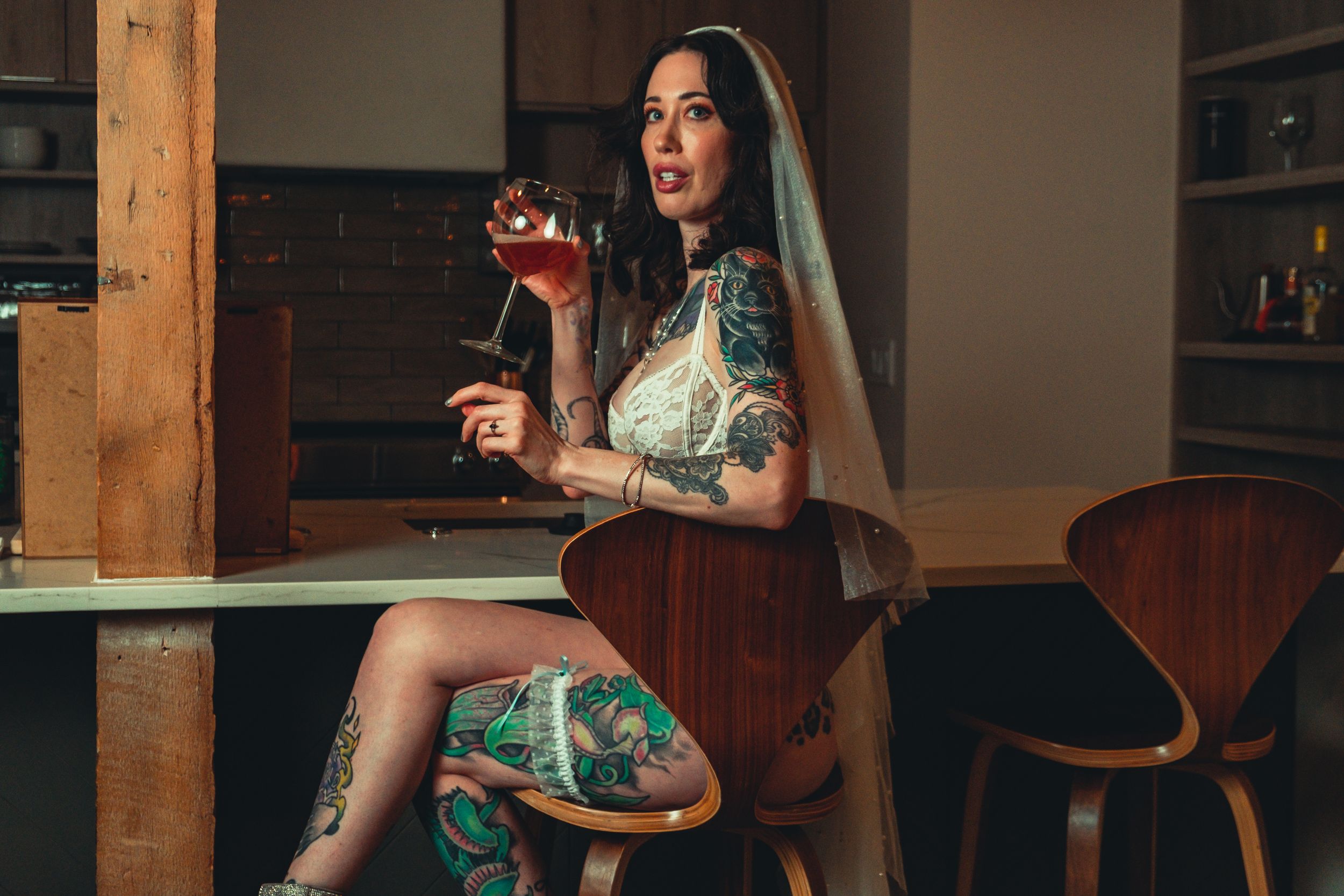 Figure in white lace sits on wooden chair holding red wine glass in moody kitchen lighting. bridal boudoir