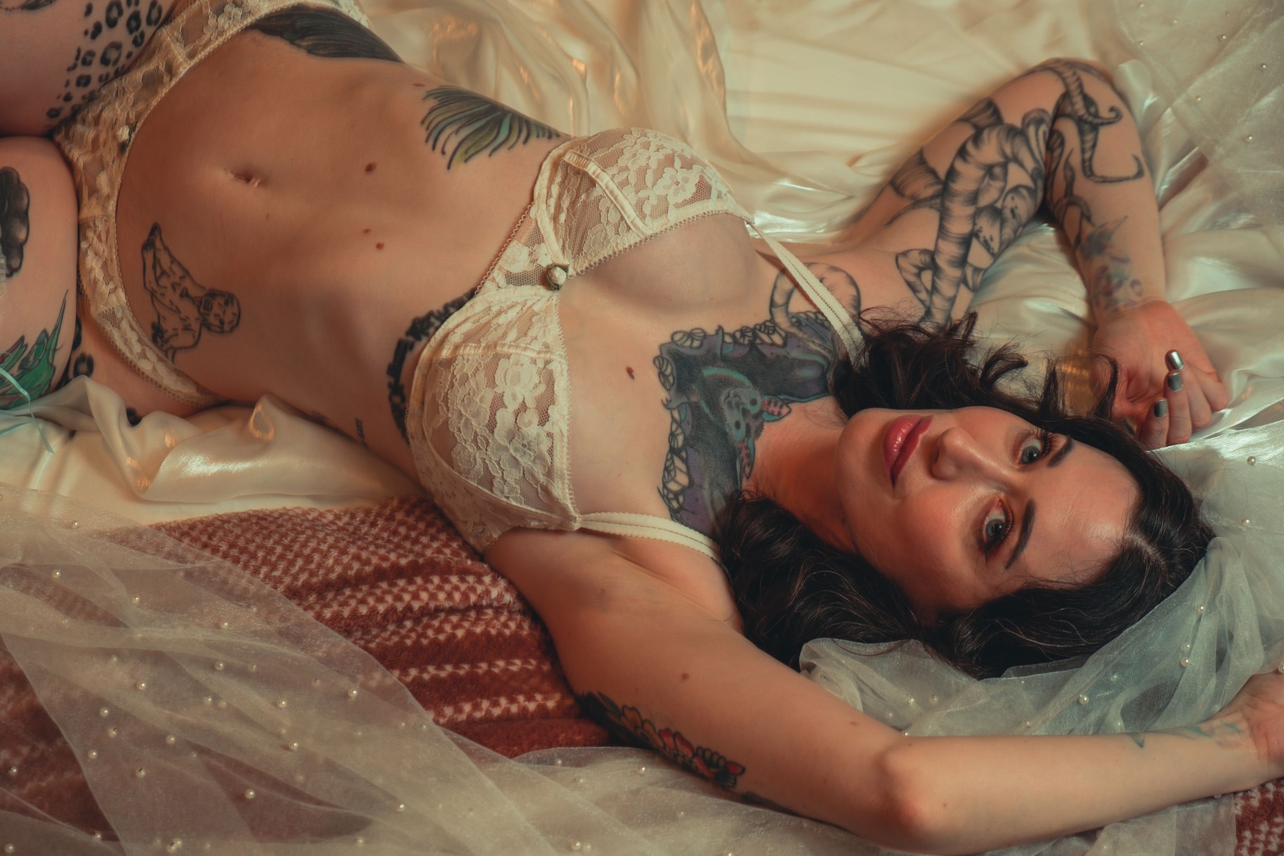 Intimate bridal boudoir photography featuring white lace lingerie and tattoos on cream textured bedding.