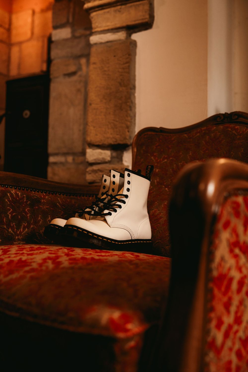White leather combat boot resting on a vintage red velvet chair in warm lighting.