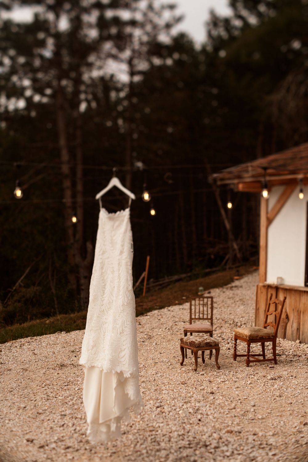 White lace wedding dress hanging outdoors with wooden chairs and string lights at dusk.