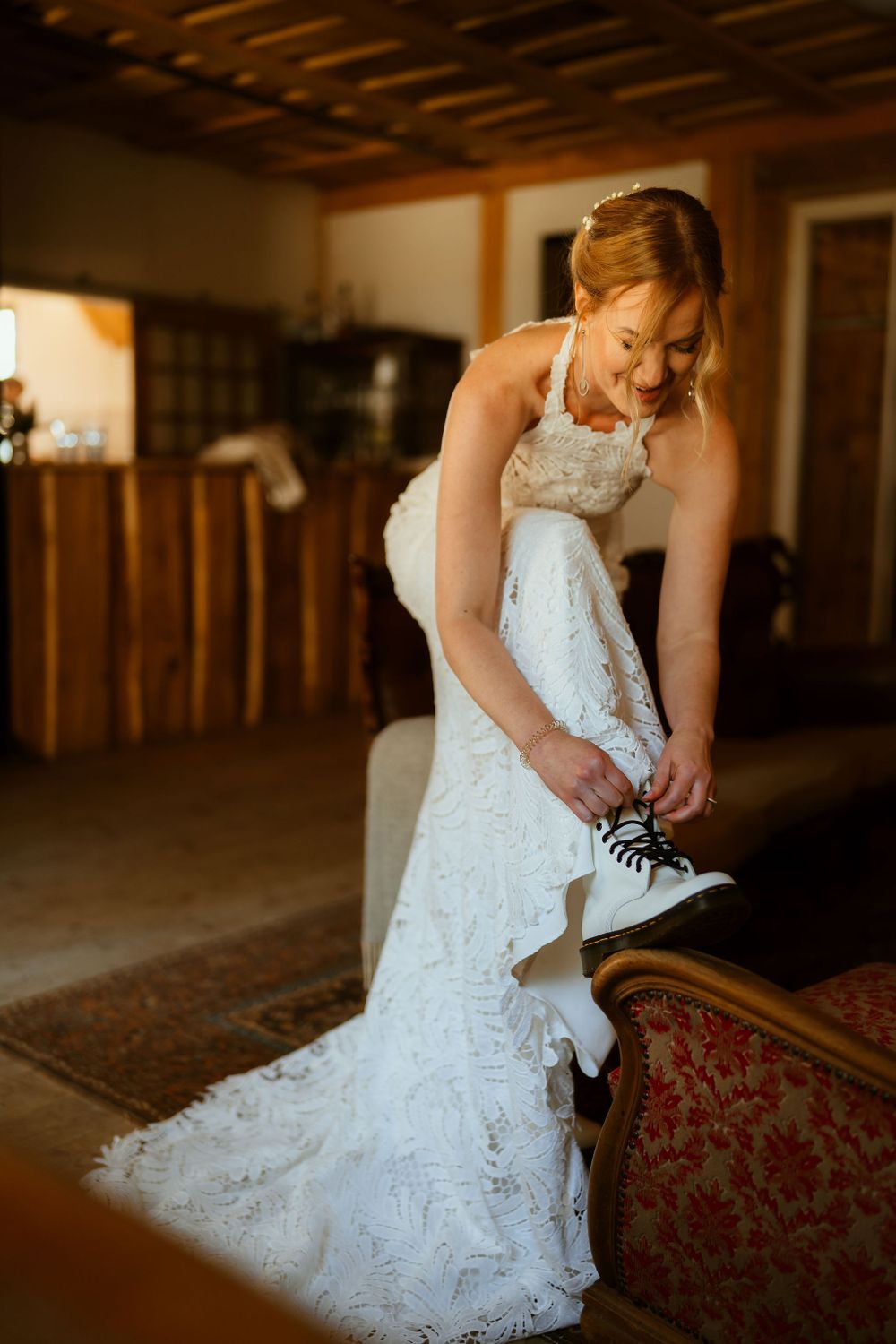 A bride in a lace wedding gown puts on white combat boots in a rustic setting.