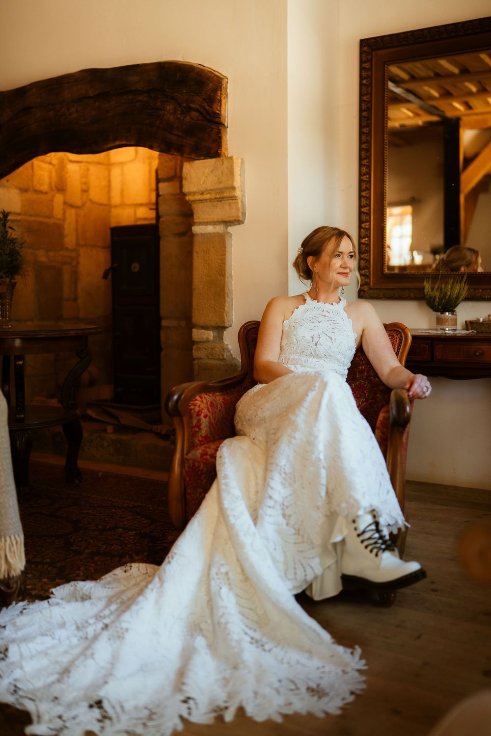 A bride wearing a lace wedding dress and boots sits elegantly in an antique chair.