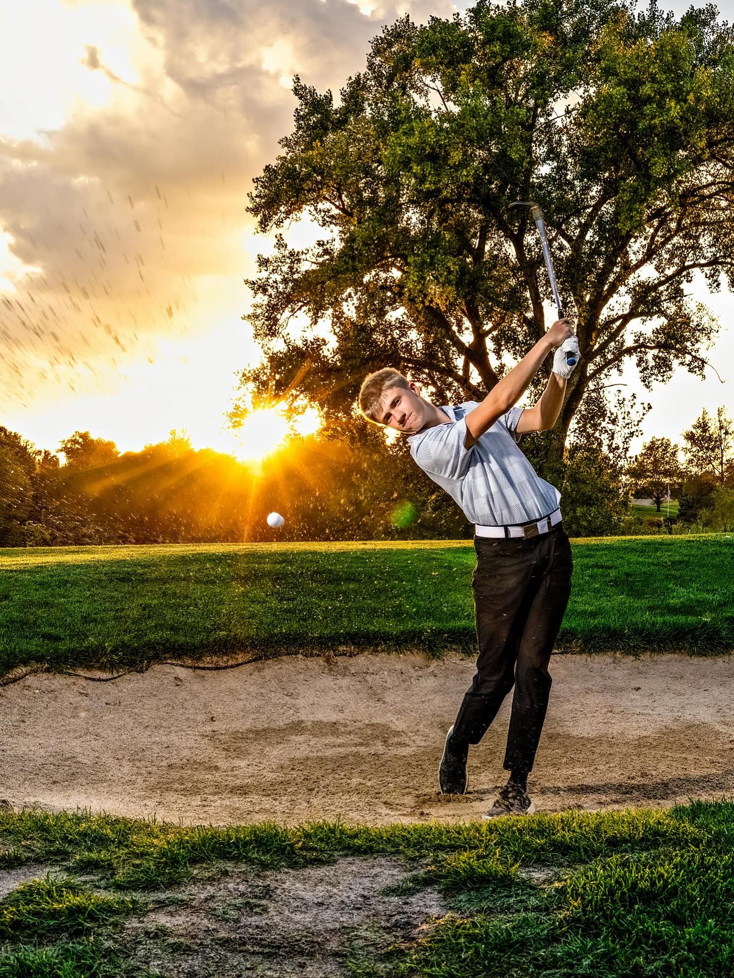 A golfer takes a swing at sunset on a scenic golf course with dramatic golden sky and silhouetted trees in the background.