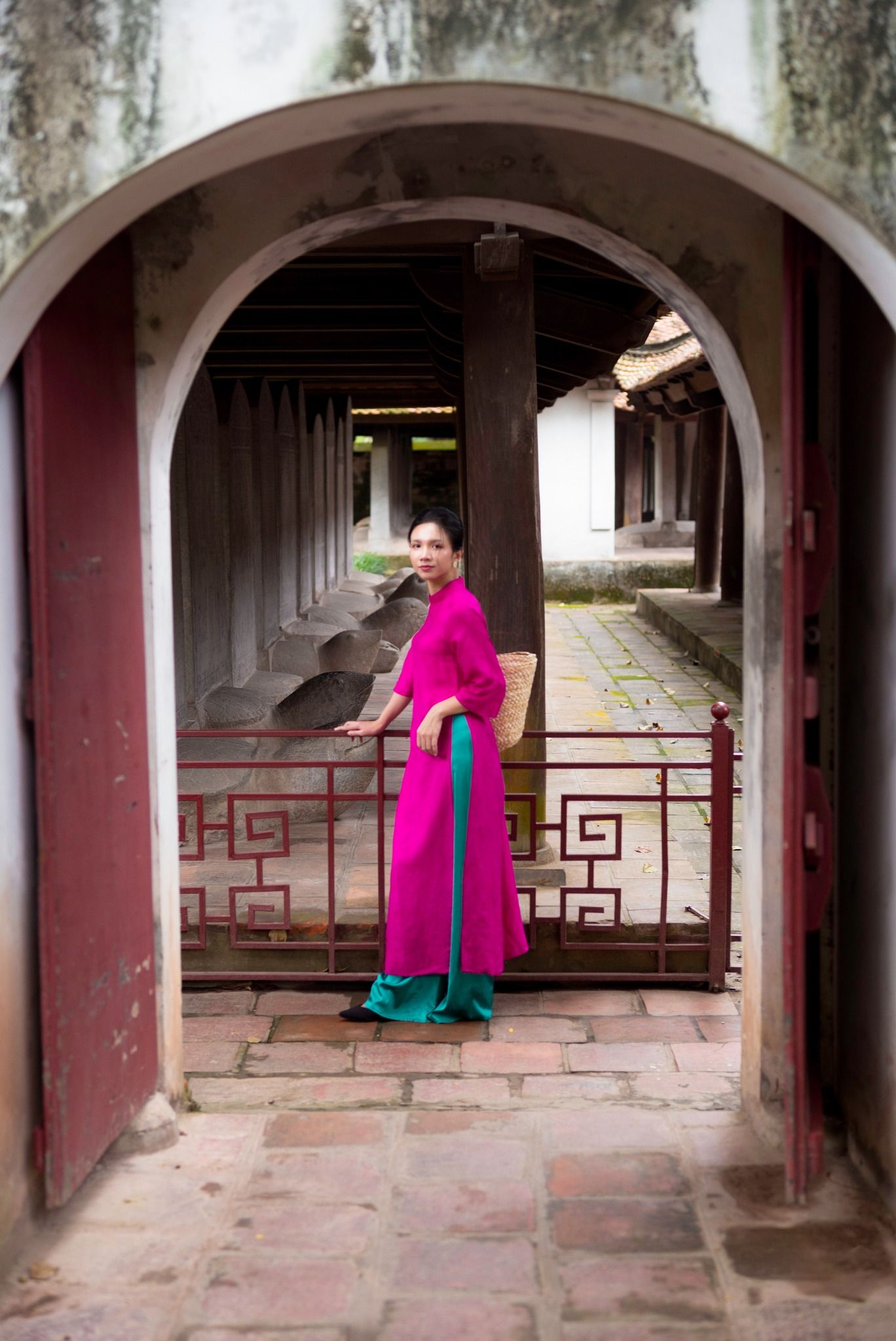Woman in pink ao dai standing at ornate archway gate of ancient Vietnamese temple courtyard.