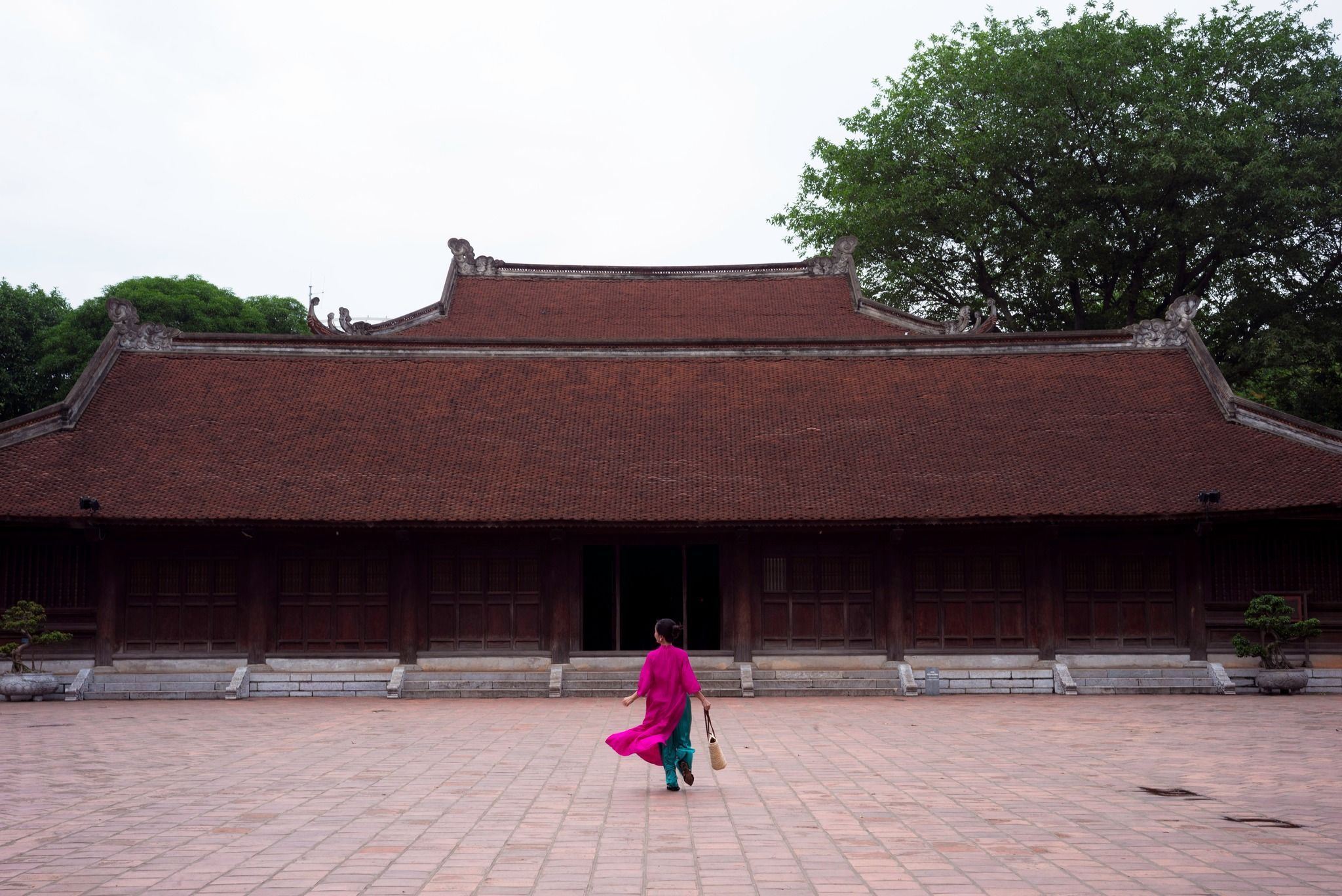 Woman in pink ao dai walking across courtyard of traditional Vietnamese wooden temple with red roof.