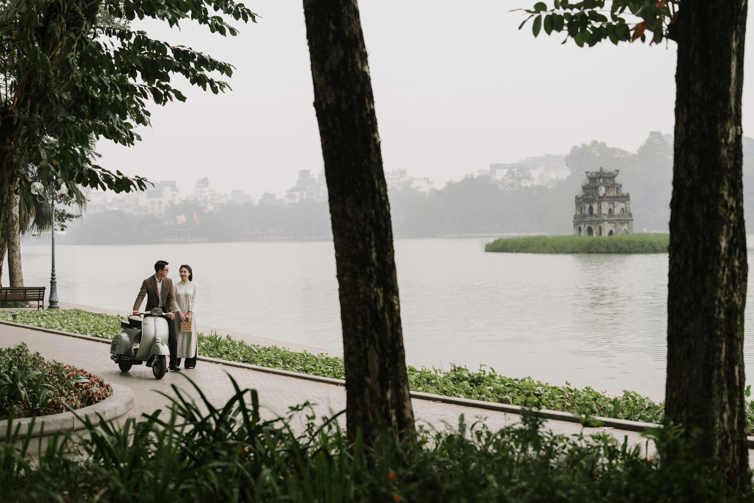 Romantic couple walking beside Hoan Kiem Lake with Turtle Tower visible through morning mist, Hanoi.