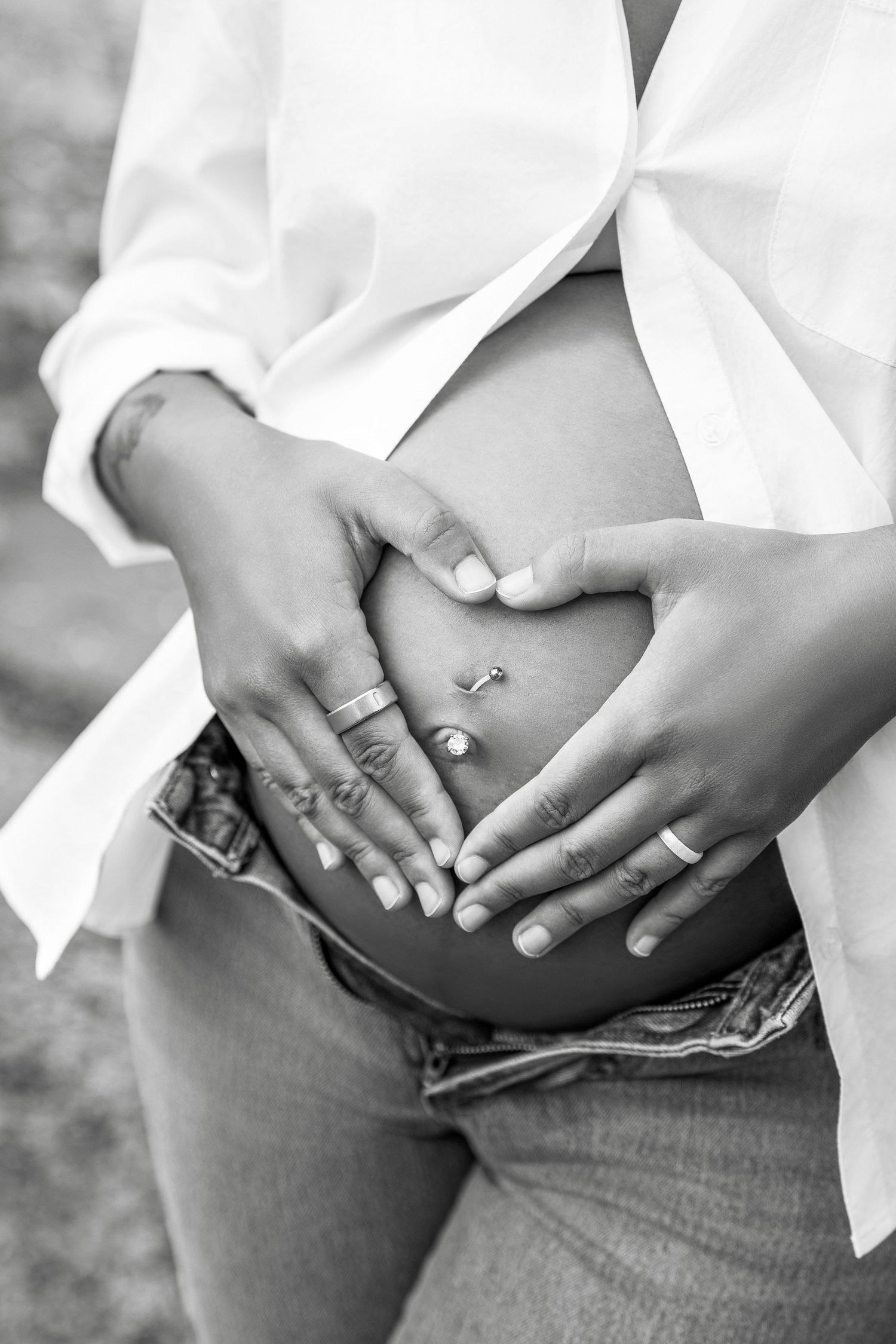 Black and white photo of hands forming heart shape on pregnant belly wearing white shirt and jeans.