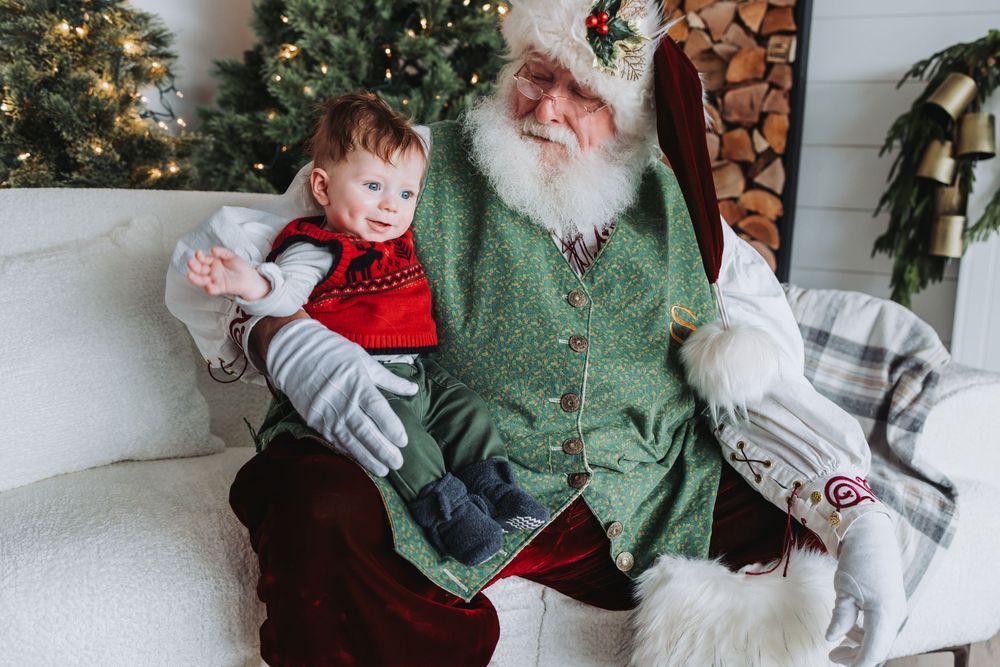 A festive holiday scene with a traditional Santa Claus in a green vest sitting on a white couch next to a small child.