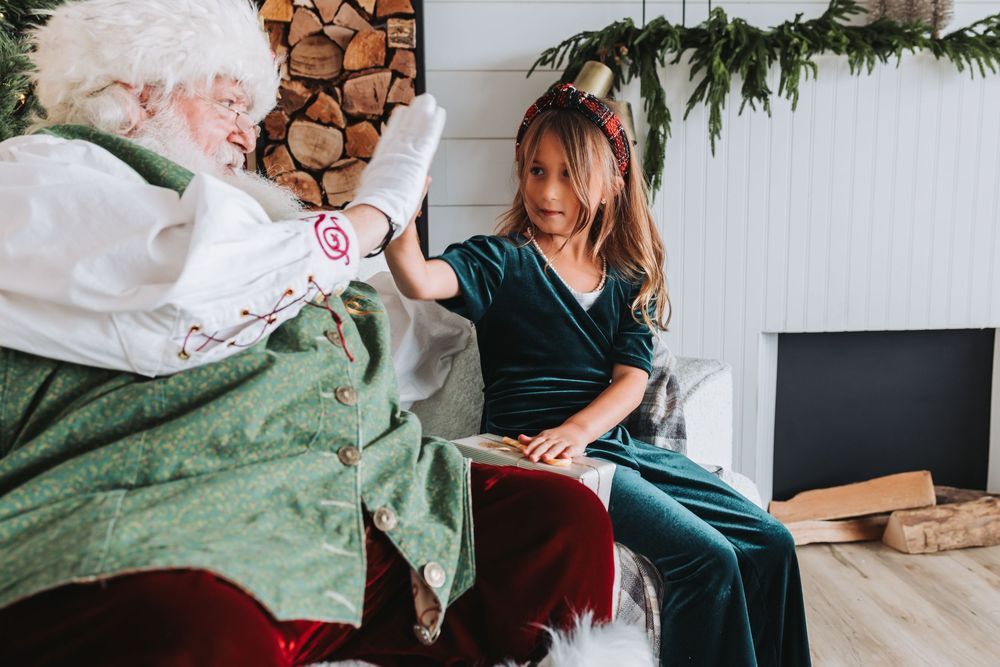 A festive holiday scene with Santa Claus in a green vest meeting with someone in a cozy indoor setting.
