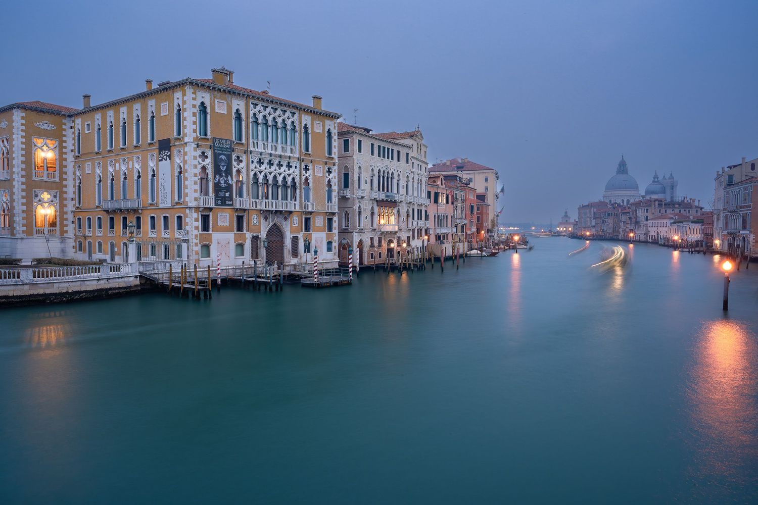 Venice's Grand Canal at dusk with historic buildings and glowing lights reflecting on calm waters.