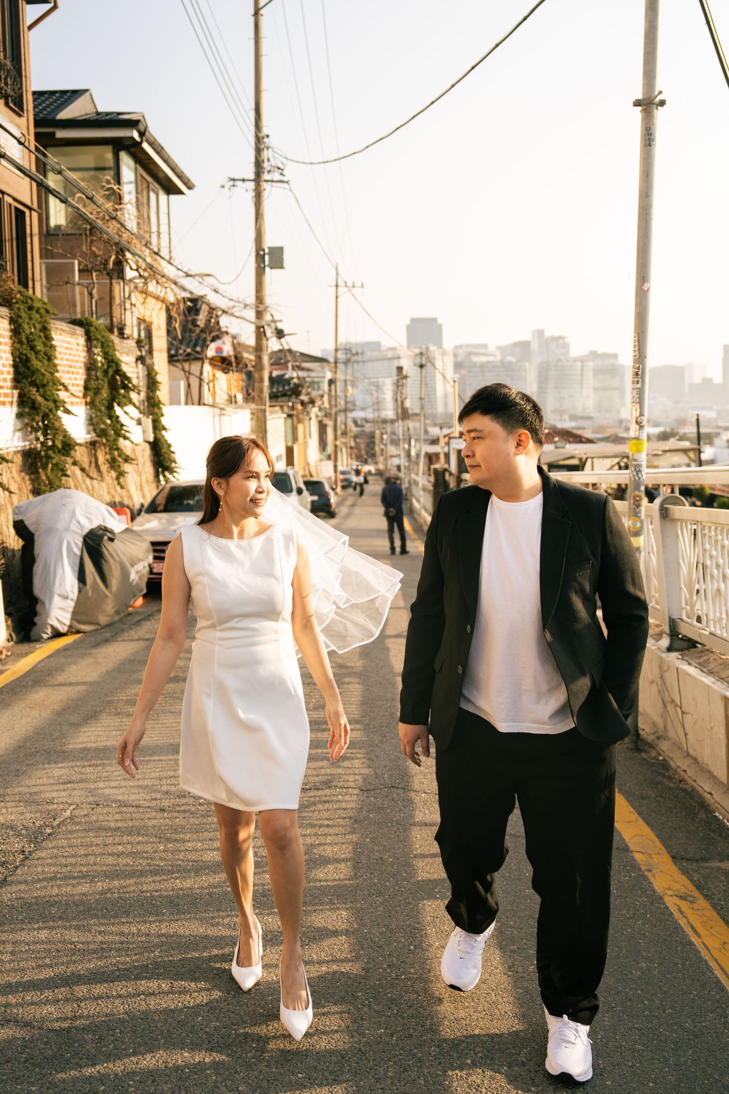 A couple in black and white attire walks hand in hand along a sunlit urban street during golden hour.