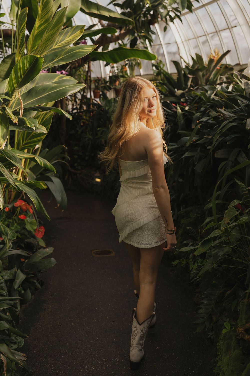 A person in a white dress poses among lush tropical plants and foliage in a greenhouse setting.