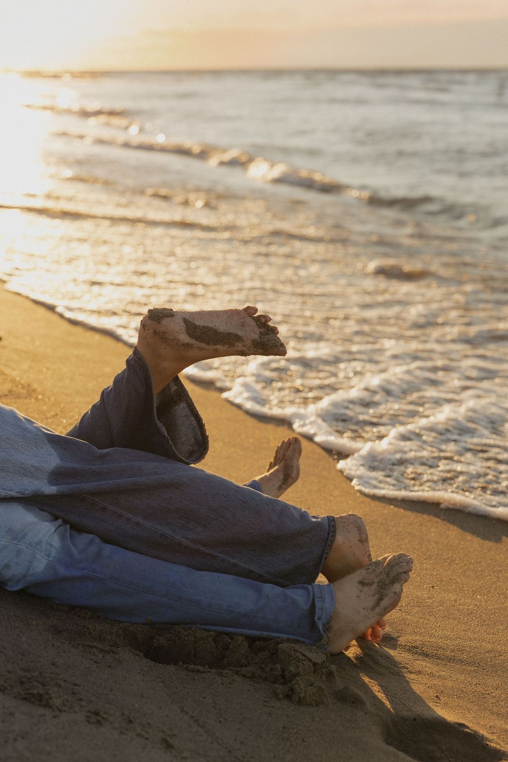 Relaxed feet in jeans resting on sandy beach at sunset with gentle waves rolling in.
