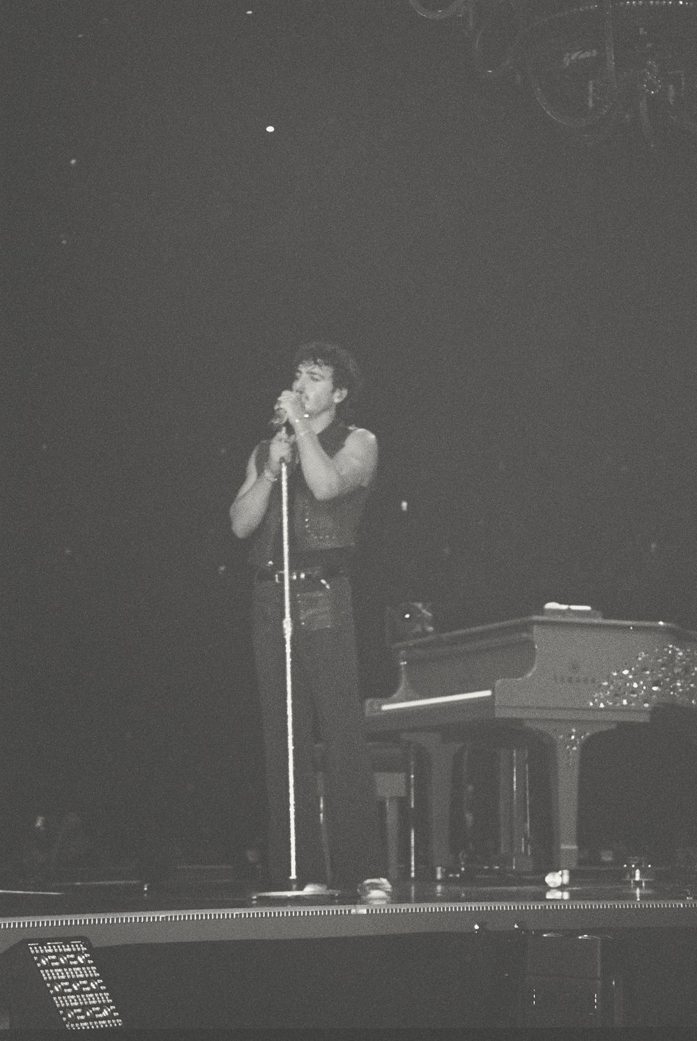 Black and white concert photo showing a performer on stage with a microphone next to a grand piano in low lighting.