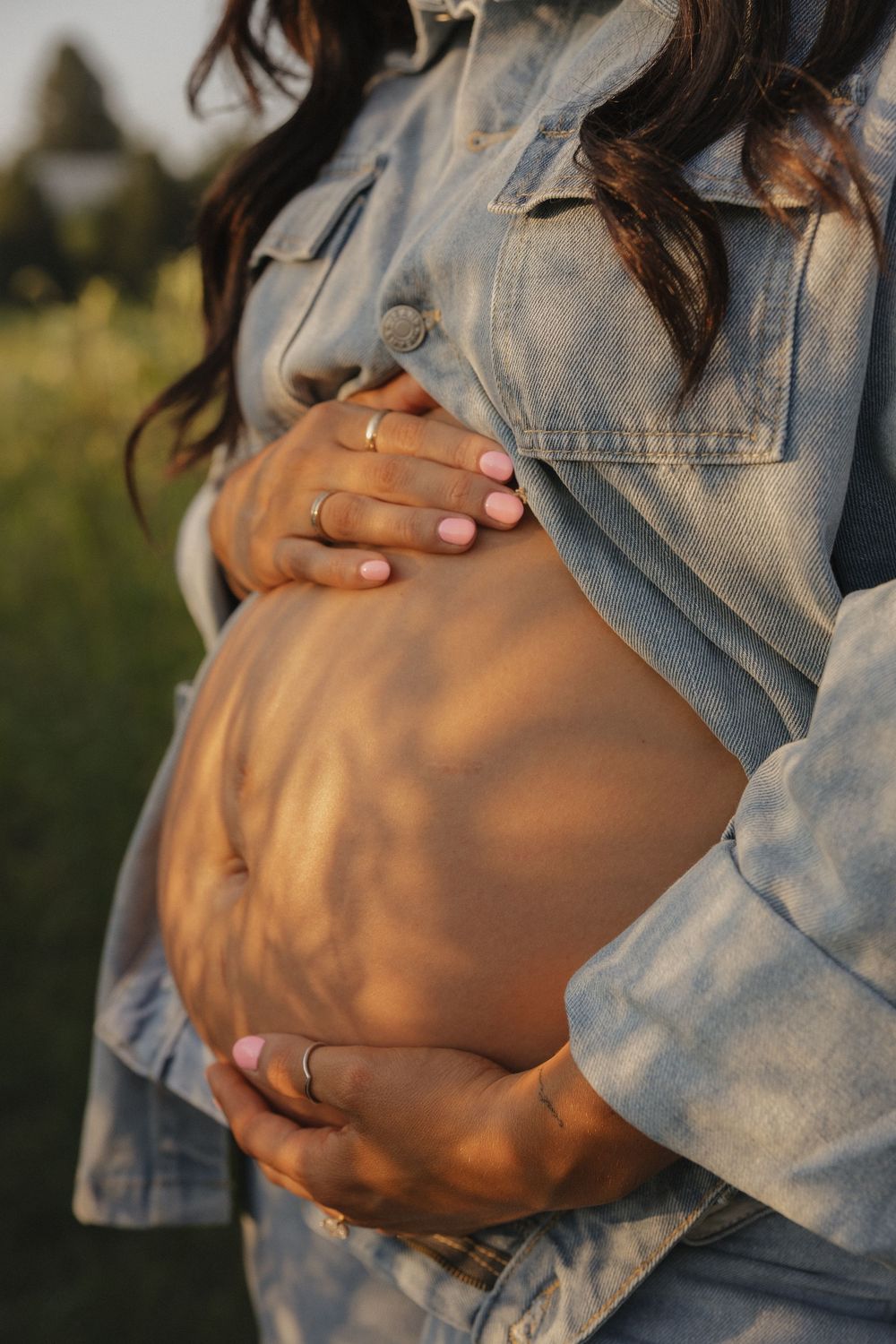 Close-up of pregnant belly being cradled by hands wearing wedding rings and a denim jacket in warm sunset light.