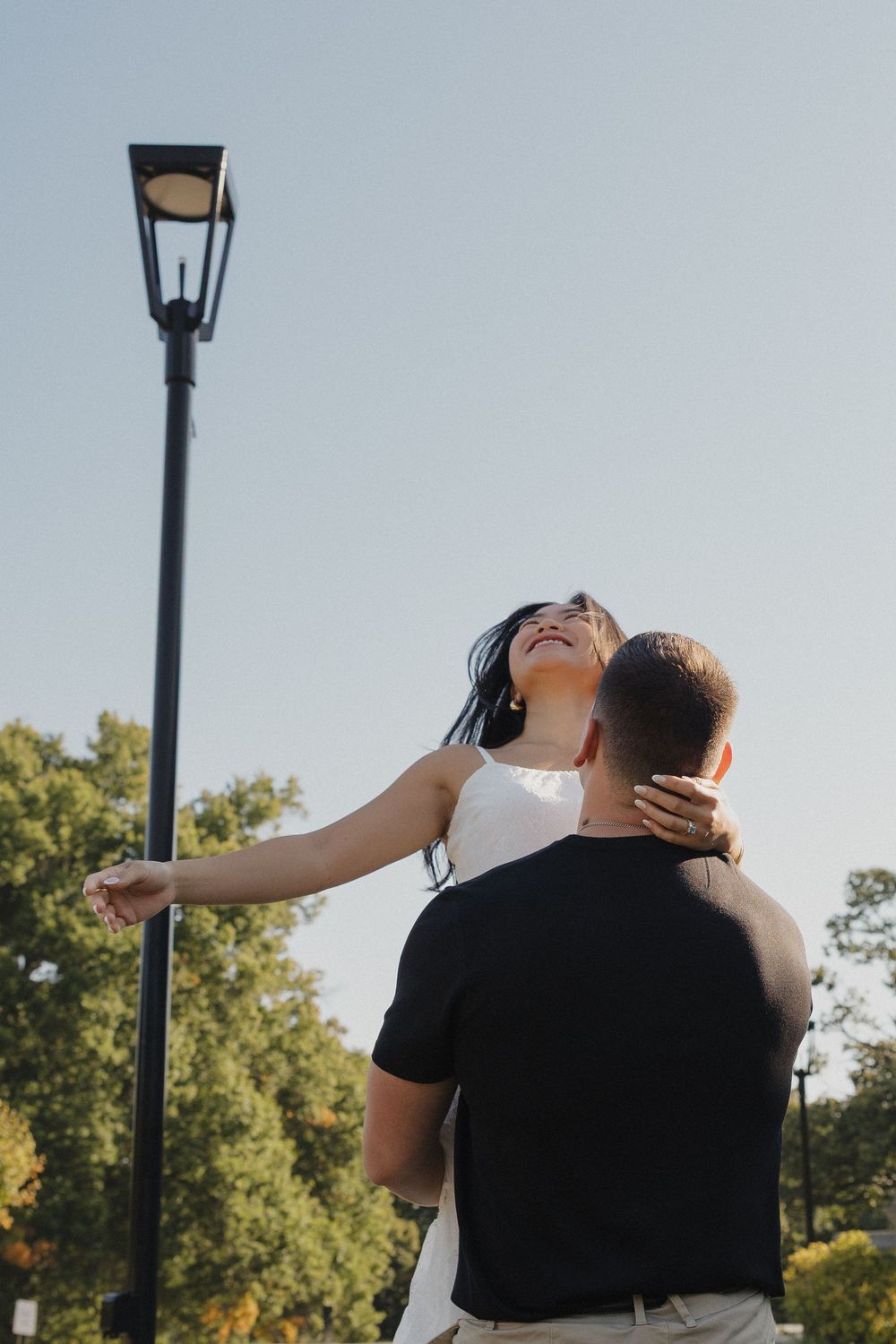 A romantic couple embraces outdoors near a vintage lamppost, with sunlit trees in the background.
