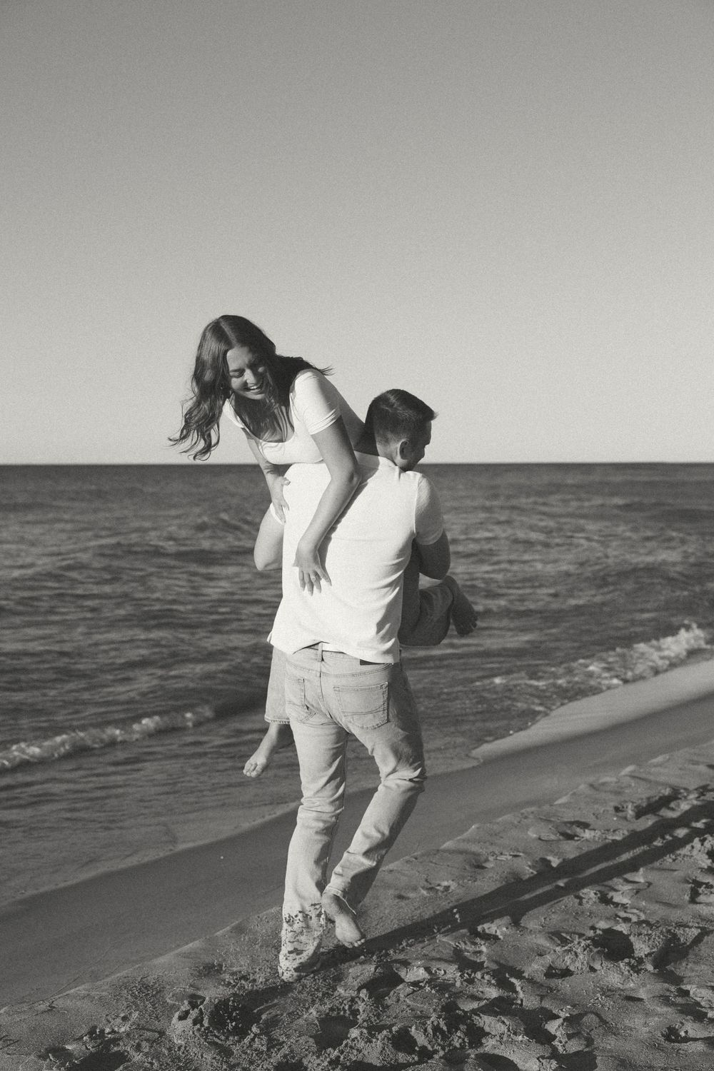 A couple in white clothing playfully dances and embraces on a beach at sunset in black and white photography.