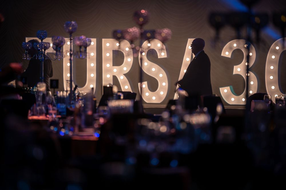 Illuminated marquee letters spelling 'FIRST 3K' with blue lighting and stage equipment visible in the background.