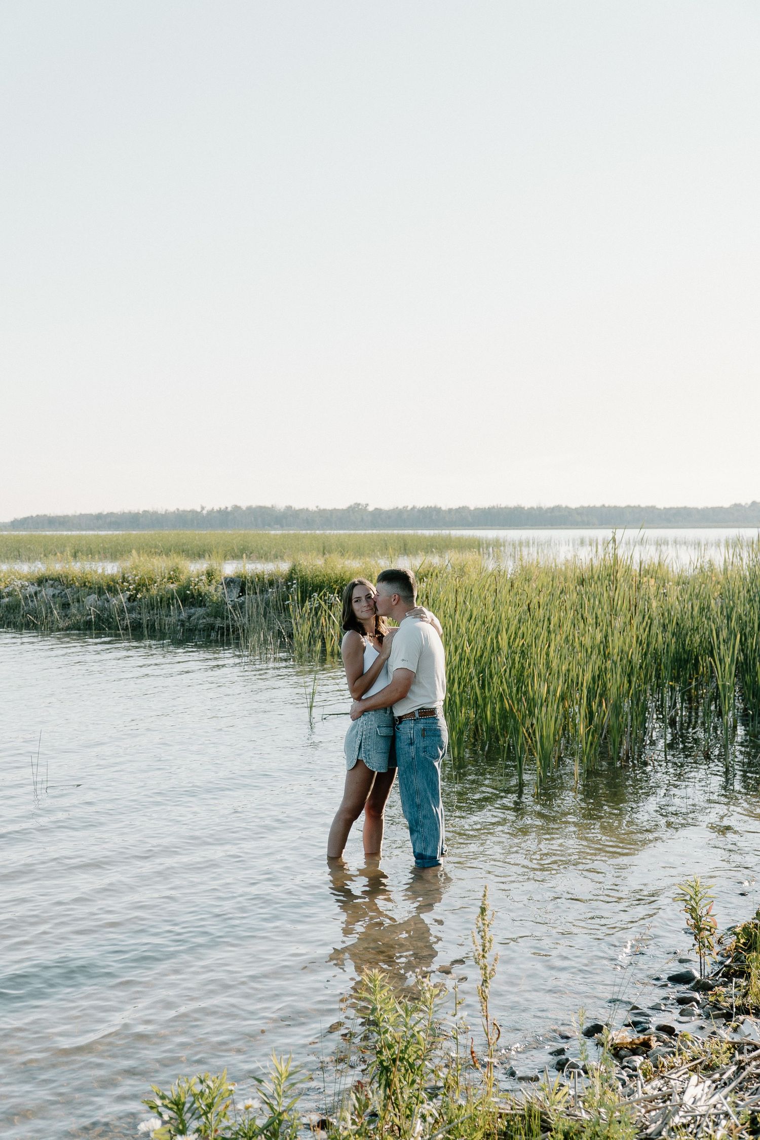 A couple shares a romantic moment along the shoreline of a serene lake with tall grass and reeds at sunset.