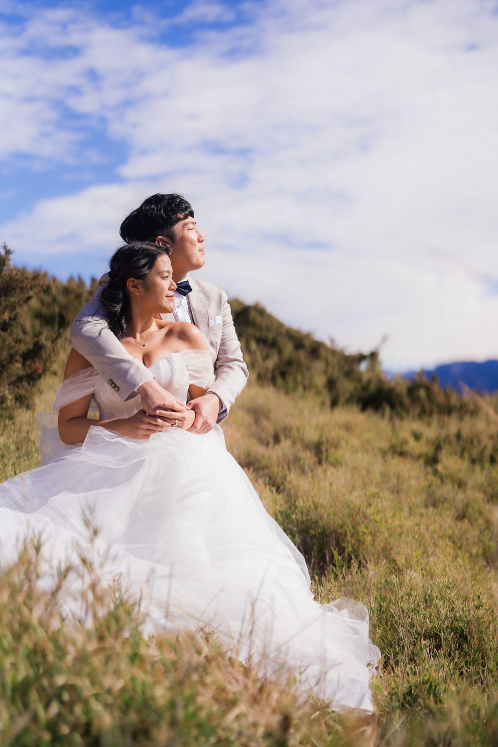 A couple in wedding attire embraces while looking into the distance on a scenic hilltop with blue skies and golden grass hear Hehuanshan.
