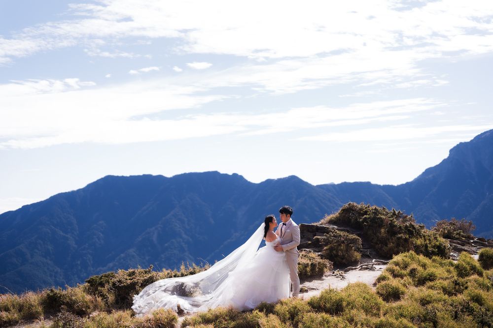A wedding couple poses on a mountain ridge with dramatic blue mountain peaks and cloudy skies in the background at Hehuanshan, Taiwan.