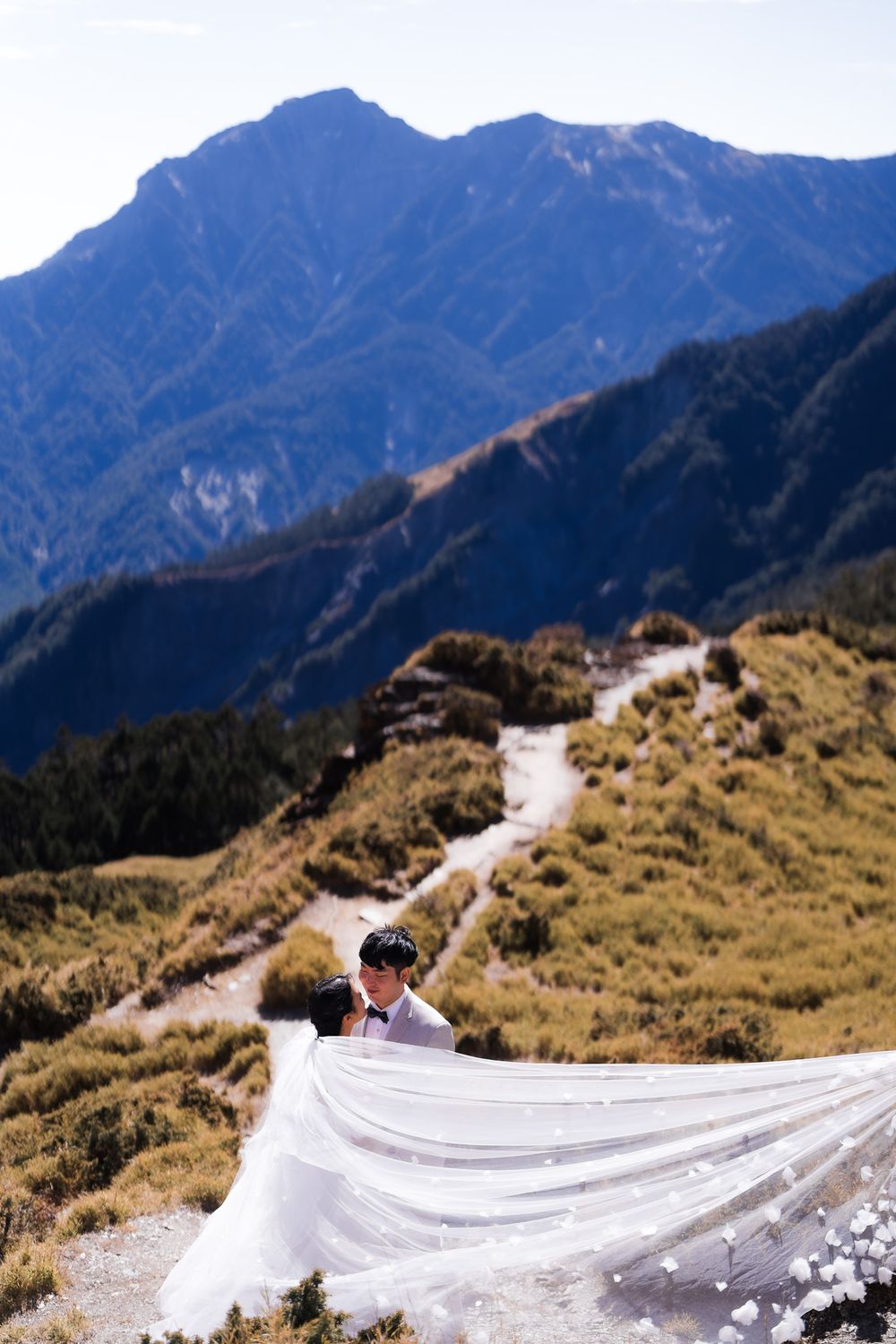 A beautiful flowing white wedding dress train cascades across a mountain top scenery with majestic peaks in the background on a sunny day