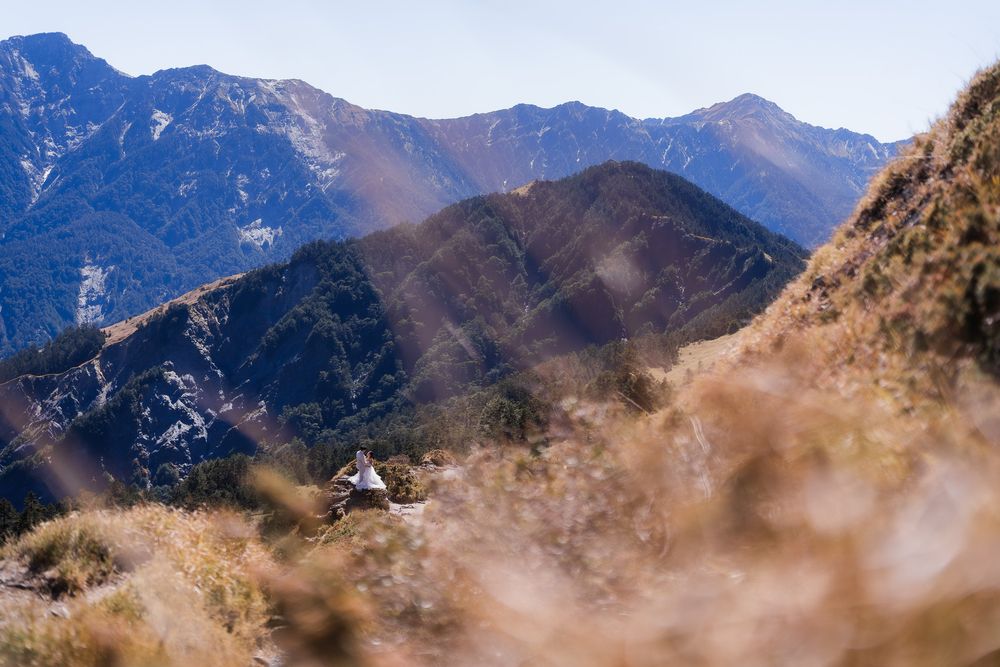 Panoramic view of a bide and groom amongst rugged mountain peaks with misty clouds rolling across the rocky landscape during daytime.