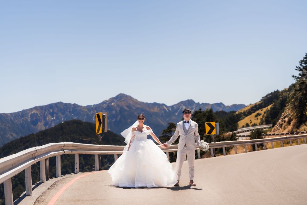 A scenic view of a bride and groom holding hands standing on a winding mountaintop road with panoramic mountain views in the background.