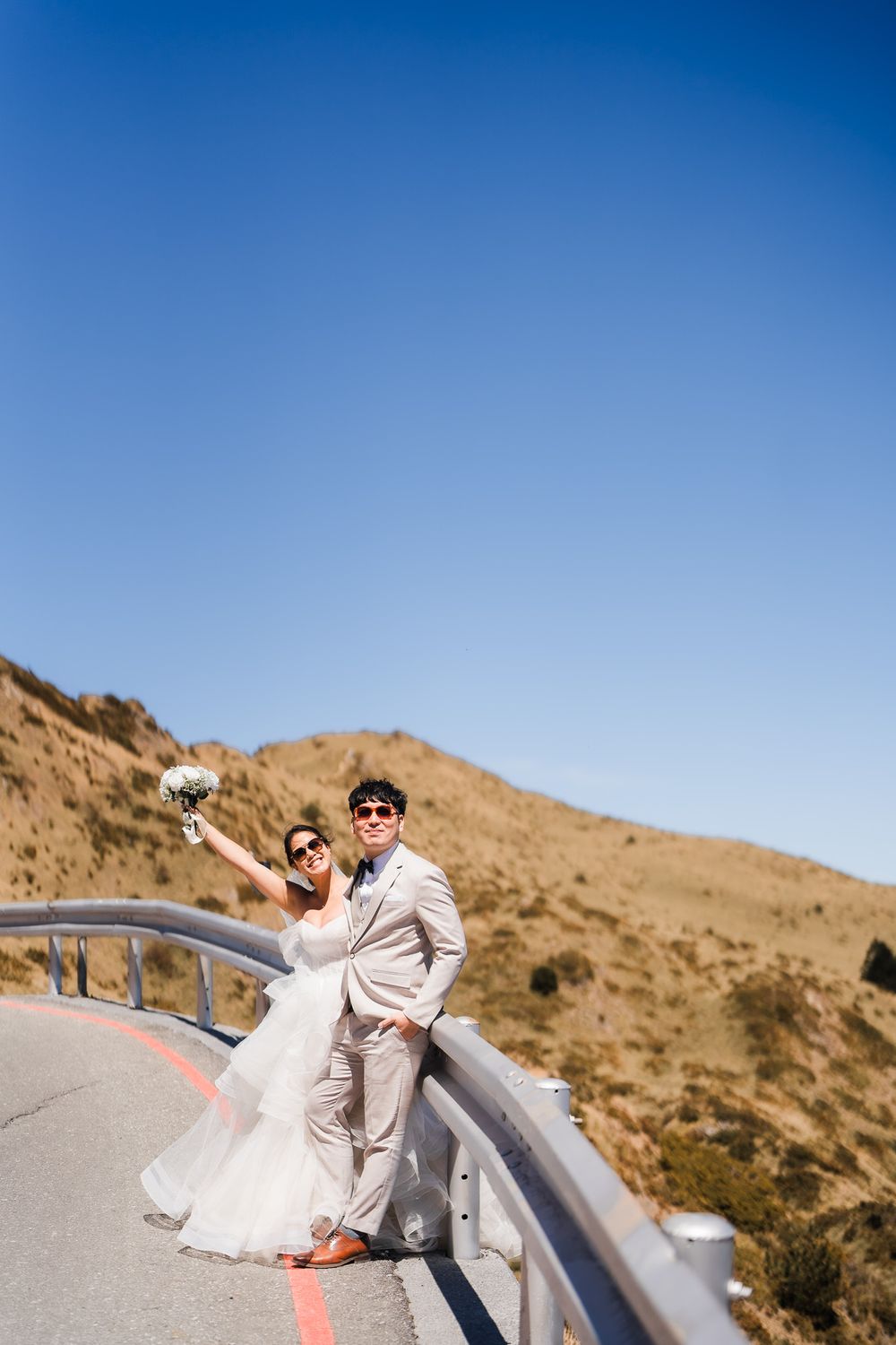 A couple in light-colored wedding attire poses playfully on a mountain road with scenic sunny hills in the background at Taiwan's Hehuanshan