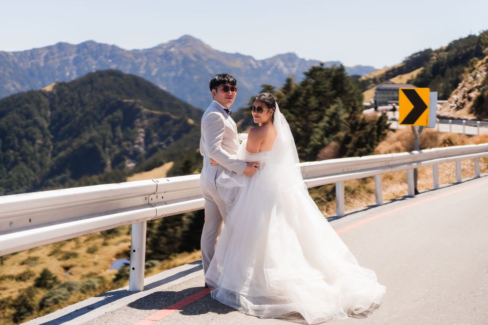 A fashionable couple in wedding attire sits on a roadside railing overlooking scenic mountain views on their special day.