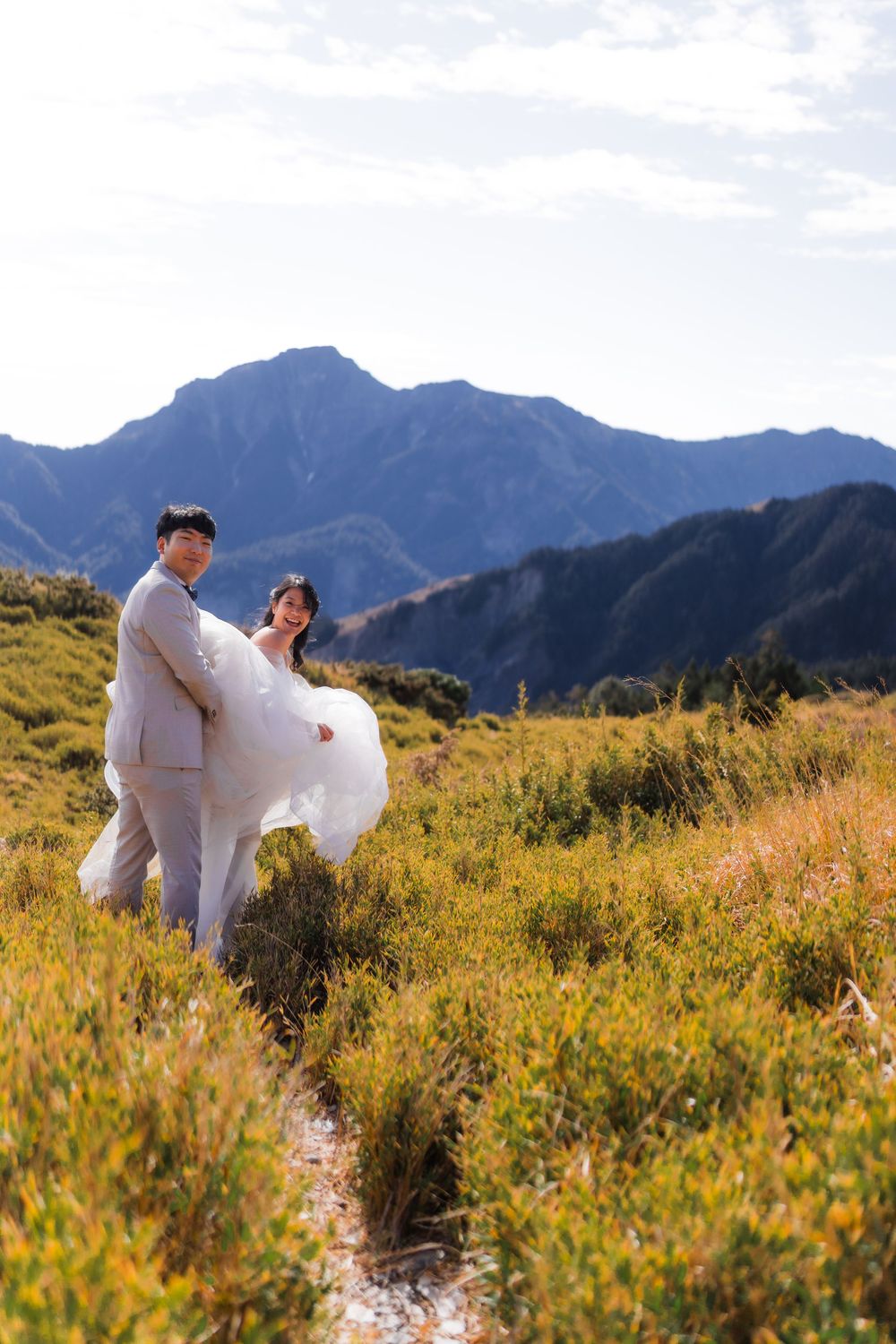 A bride and groom playfully poses in a mountain meadow filled with yellow wildflowers on a sunny day.