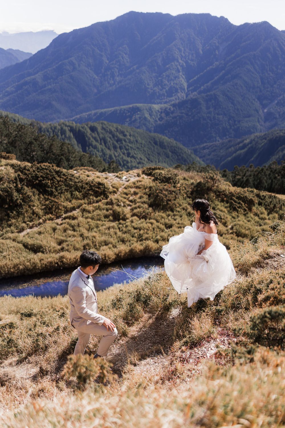 A bride and groom walk dramatically along the hillside on a mountain hillside with stunning peaks in the background.