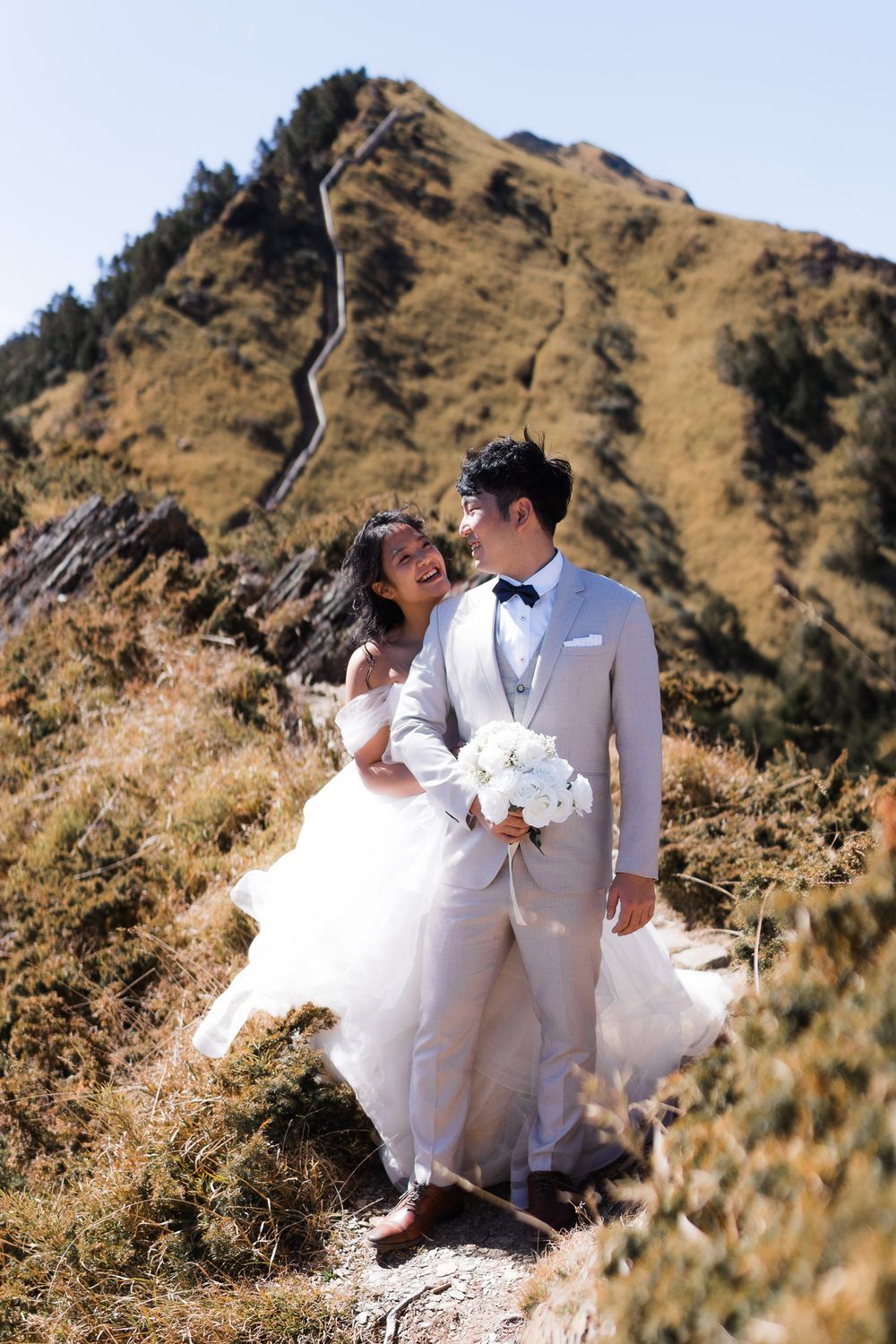 A bride embraces her groom while walking along a mountainside path leading up to a rugged peak on a sunny day at Heshuanshan, Taiwan.