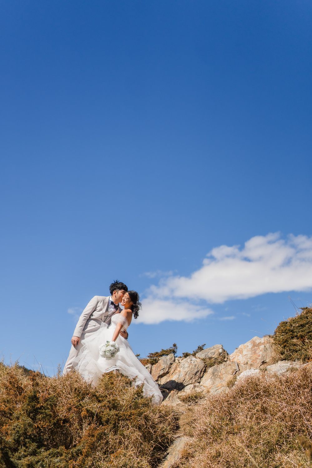 A bride and groom in white wedding attire share an intimate kiss atop a rocky hillside against a bright blue sky at Hehuanshan, Taiwan.