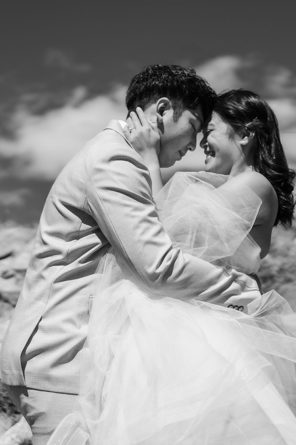 A romantic black and white photo showing a couple in white wedding attire sharing an intimate embrace outdoors against a cloudy sky.