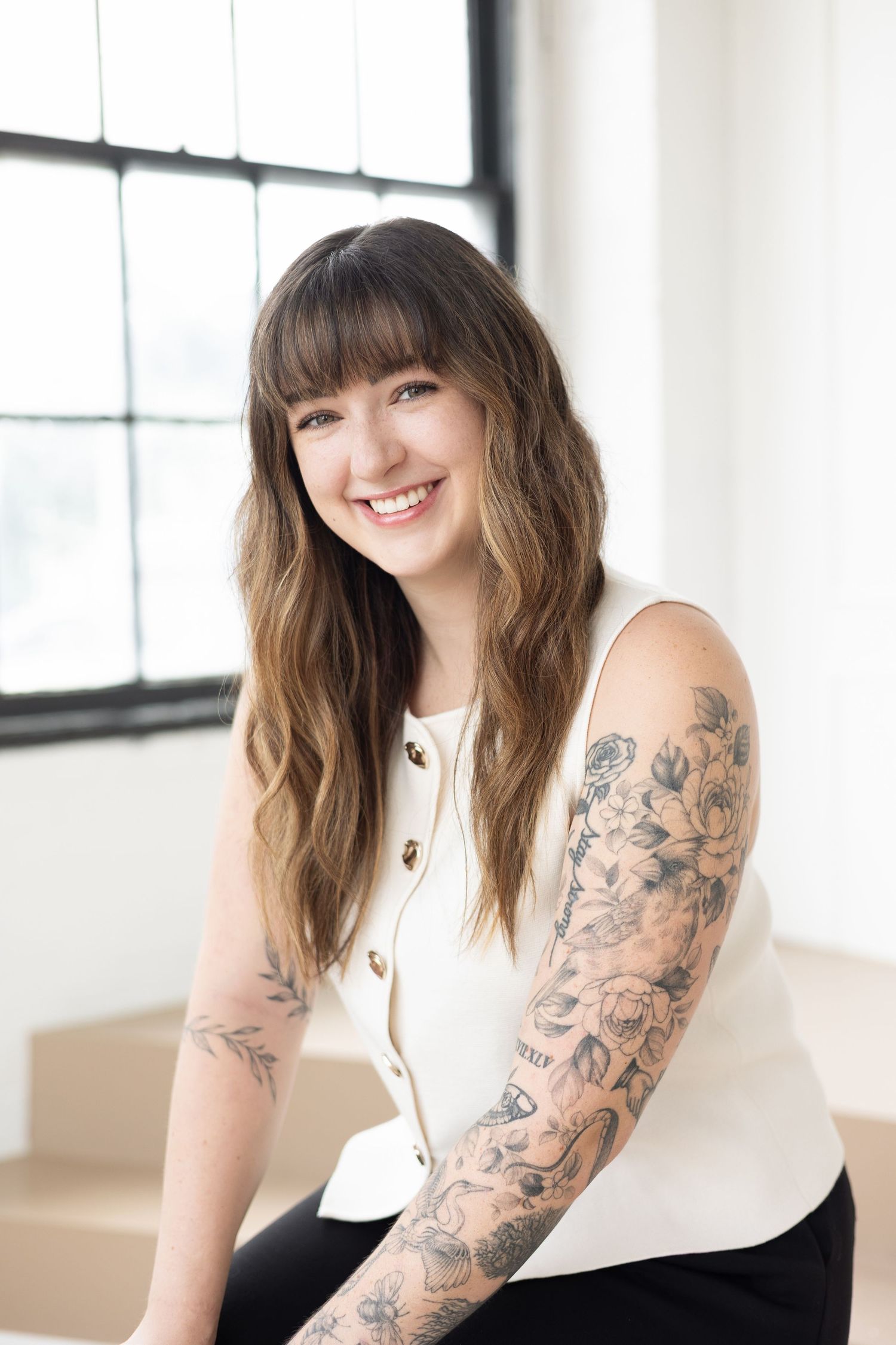 Nurse injector with warm smile wearing a white sleeveless top with gold buttons and tattoos on arms at Parkhill Studios in Cambridge.