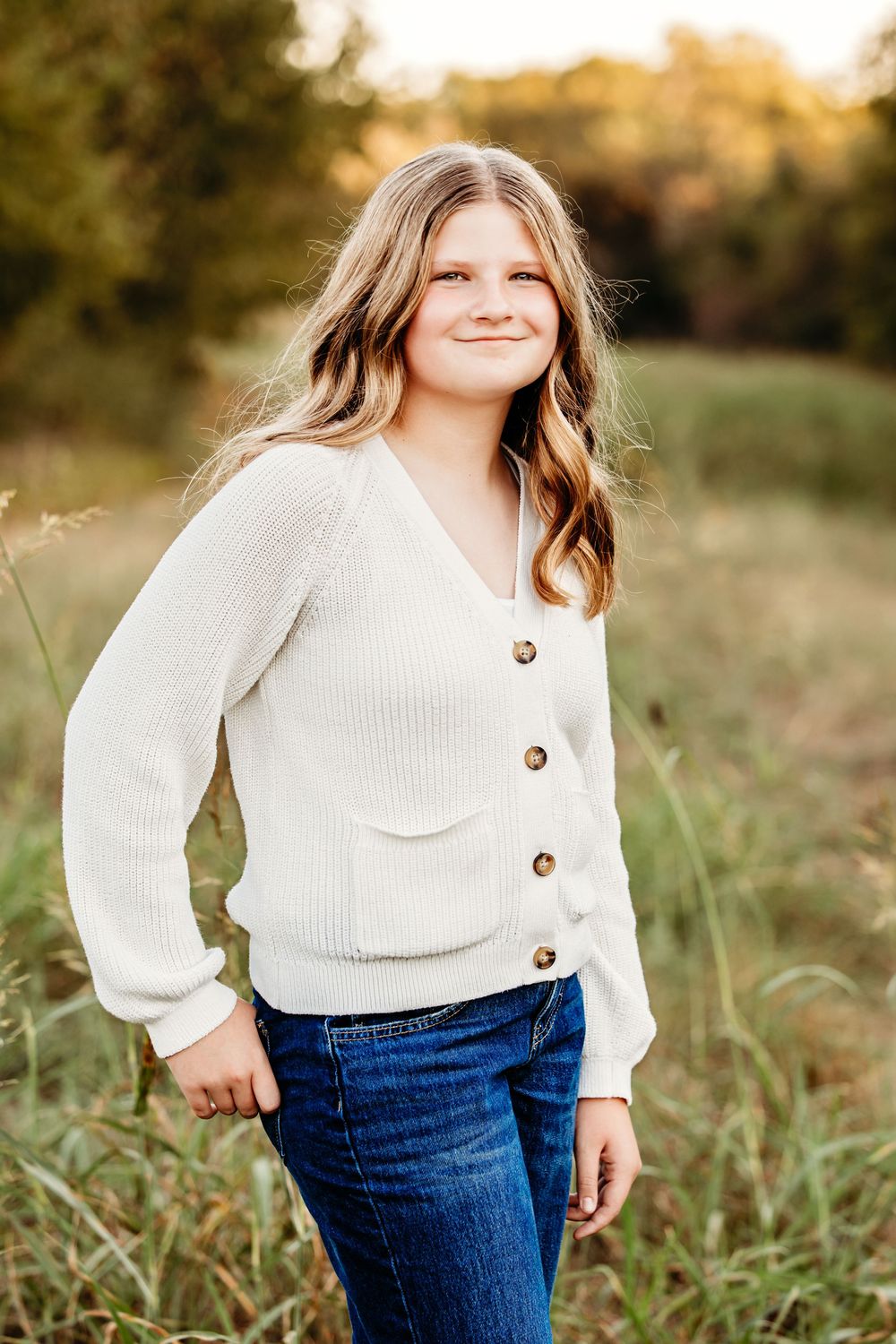 Portrait of someone wearing a cream knit cardigan and jeans standing in a sunlit field at golden hour.
